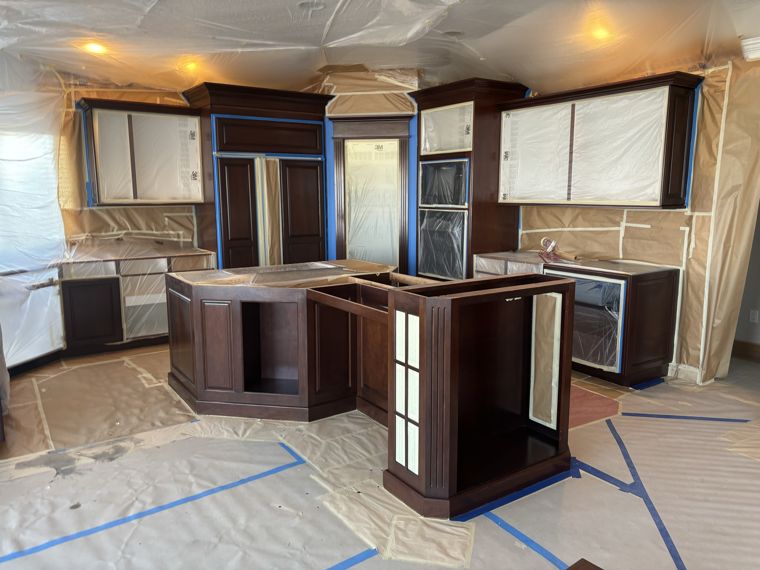 Room with dark wood cabinets and display cases being painted, covered with protective plastic and painter's tape, with plastic-lined floor and ceiling. Kitchen remodel cabinet painting in the Treasure Valley.