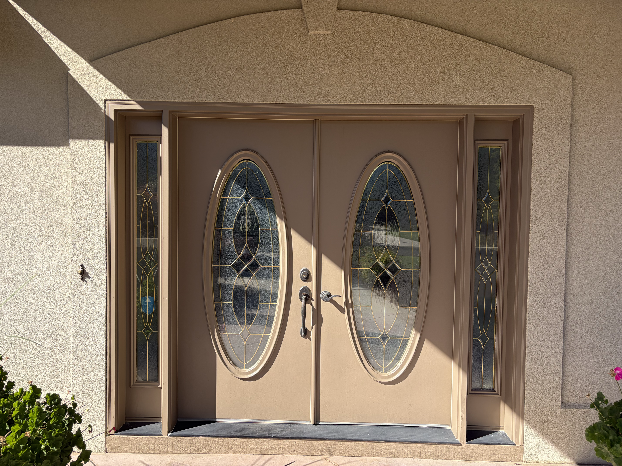 Front door with decorative glass panels and a handle, surrounded by beige walls and a small potted plant in the lower left corner. Completed exterior painting in Meridian, Idaho