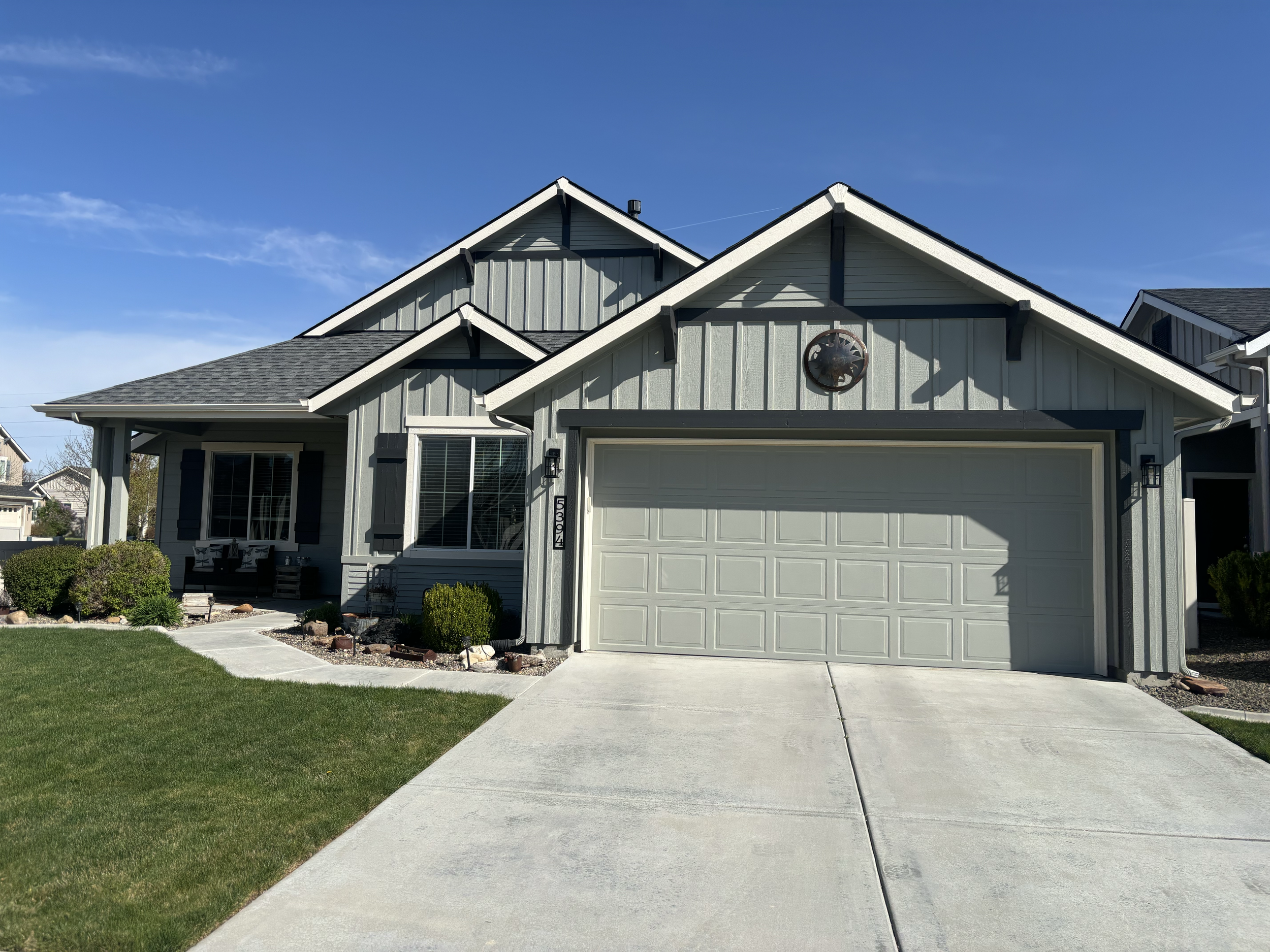 A modern two-story house with a light gray exterior, black trim, and a white garage door. The driveway is concrete, leading to a well-kept front yard with green grass and small shrubs. The house has a decorative sun-shaped element on the front façade