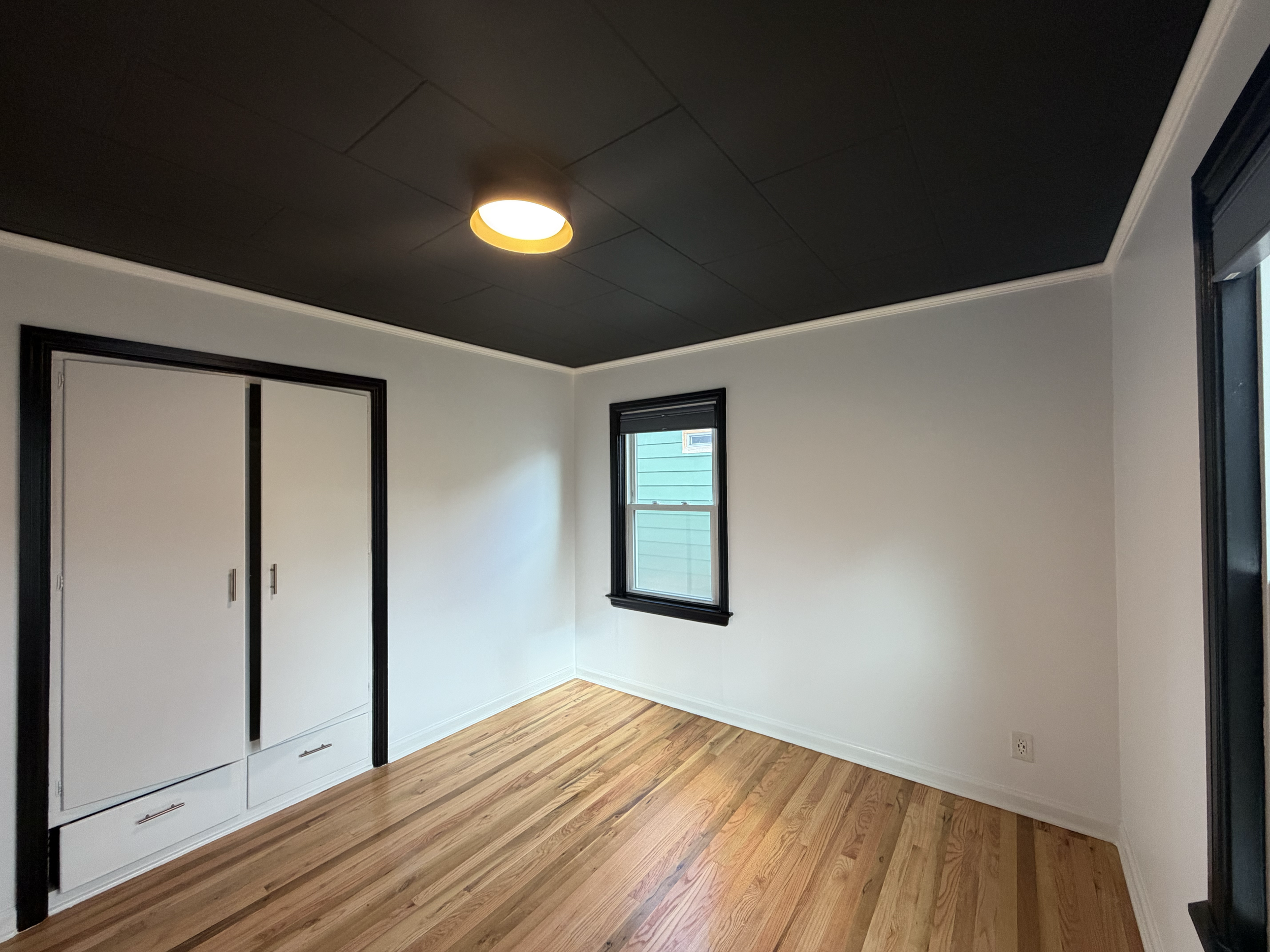 Freshly painted empty room with white walls, hardwood floor, black window trim, and black ceiling in Middleton, Idaho. There is a ceiling light and a closed sliding closet door. 