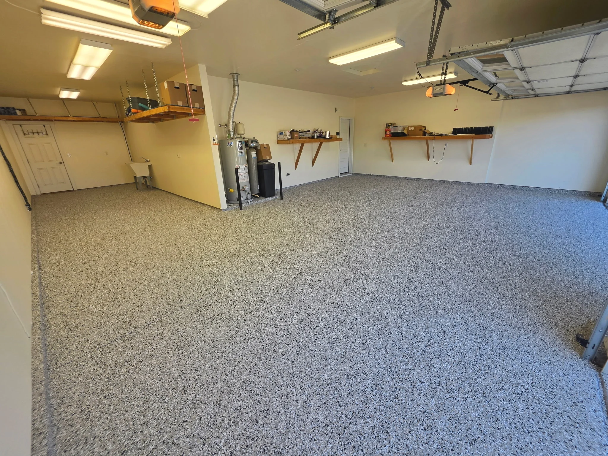 Empty garage interior with beige walls, gray speckled epoxy flooring, ceiling lights, and a partially open garage door. There are shelves with various items and a water heater near the back wall. After epoxy flooring results with flecks in it.