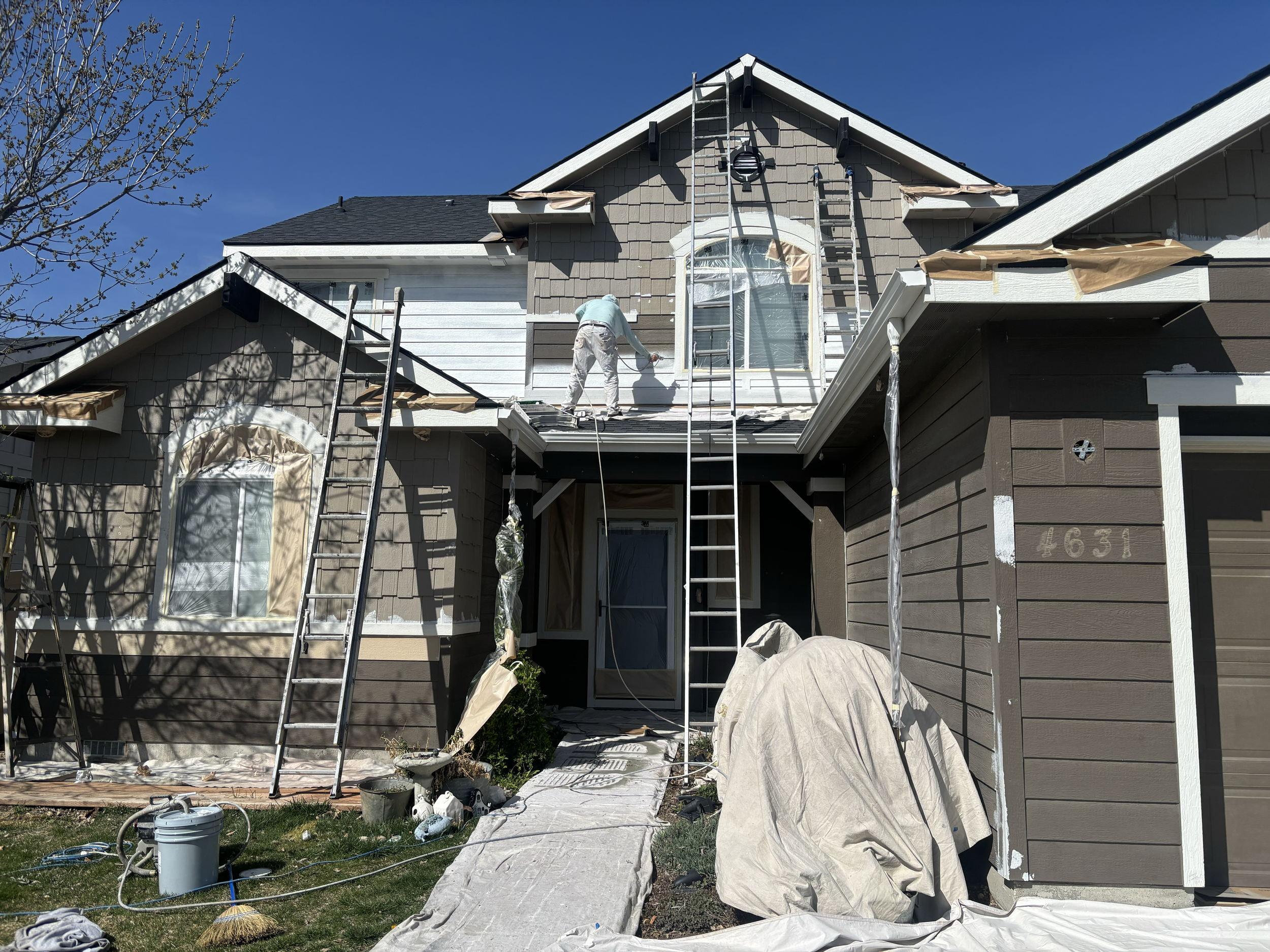 A worker painting the exterior of a house with gray and white siding. Ladders, scaffolding, and painting supplies are visible, and the house has multiple windows and a gabled roof. Multi-level exterior residential painting in Treasure Valley