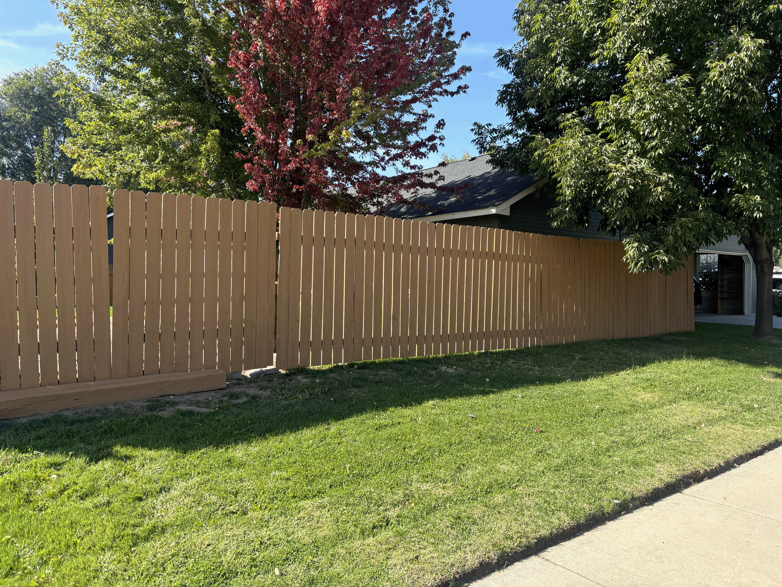 A wooden privacy fence with a suburban house and lush green trees in the background under a clear blue sky. After a fresh fence stain application by Idaho Valley Painting.