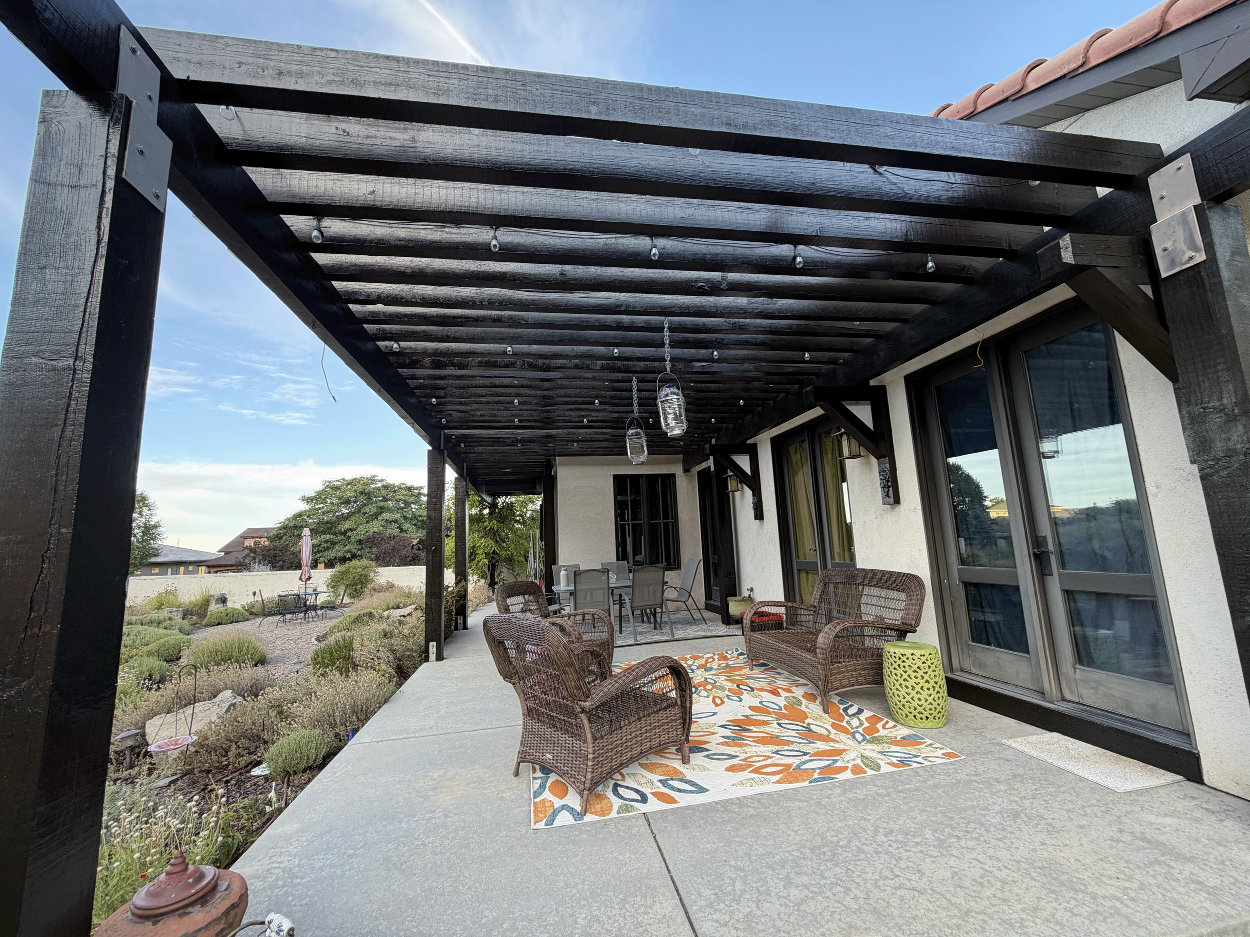 View of a backyard patio area with outdoor furniture, a decorative rug, and string lights hanging from a wooden pergola. The background shows a garden with plants and neighboring houses. After fresh dark exterior wood stain application in Boise, ID