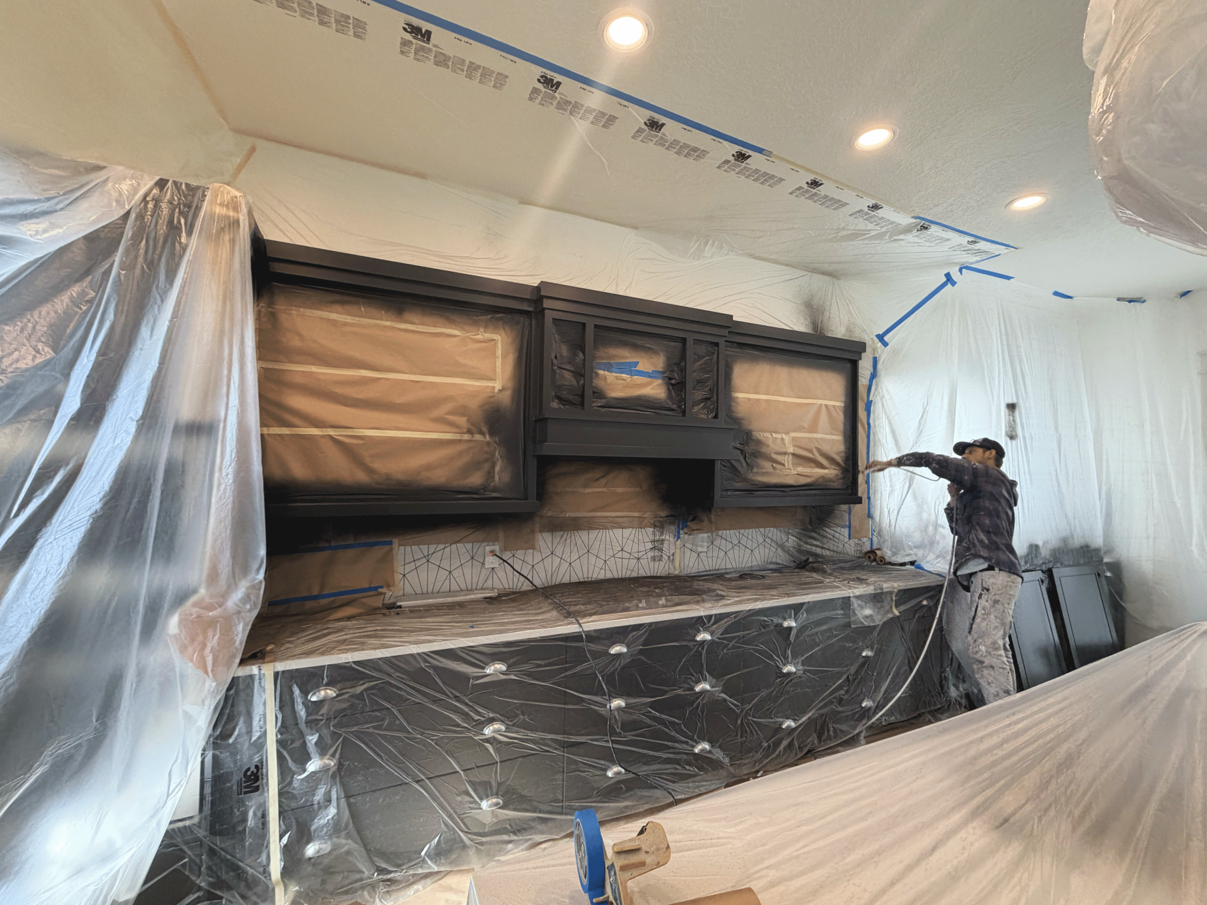 A person working on kitchen cabinet painting in a room with plastic coverings and tape on the ceiling. The room features a dark-colored cabinet or mantel, and recessed lighting in the ceiling.