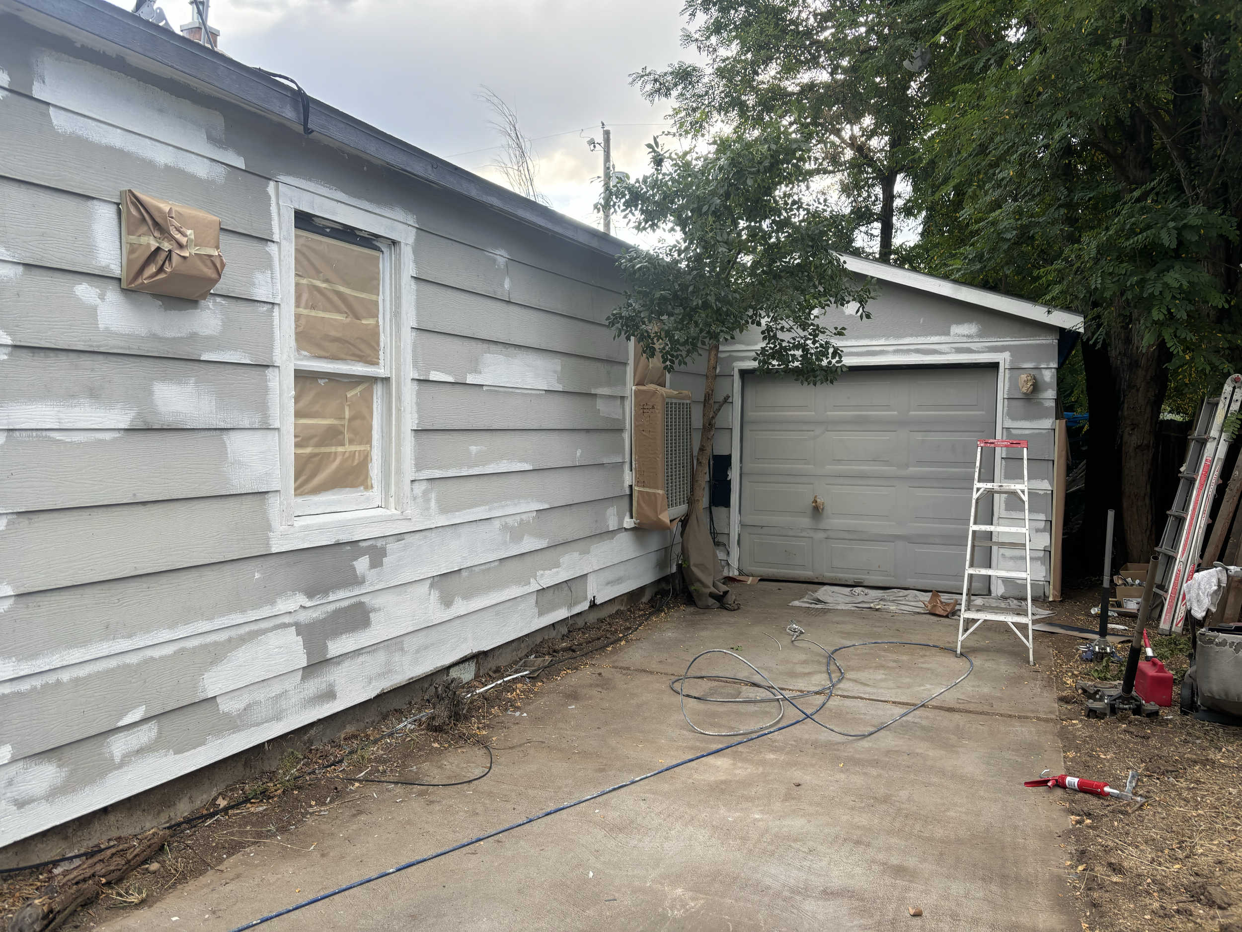 Exterior of a house undergoing painting, with the siding partially painted white and some areas taped off. There are painting supplies and ladders visible in the yard, along with a tree and a garage door. Exterior repaired before painting in Boise