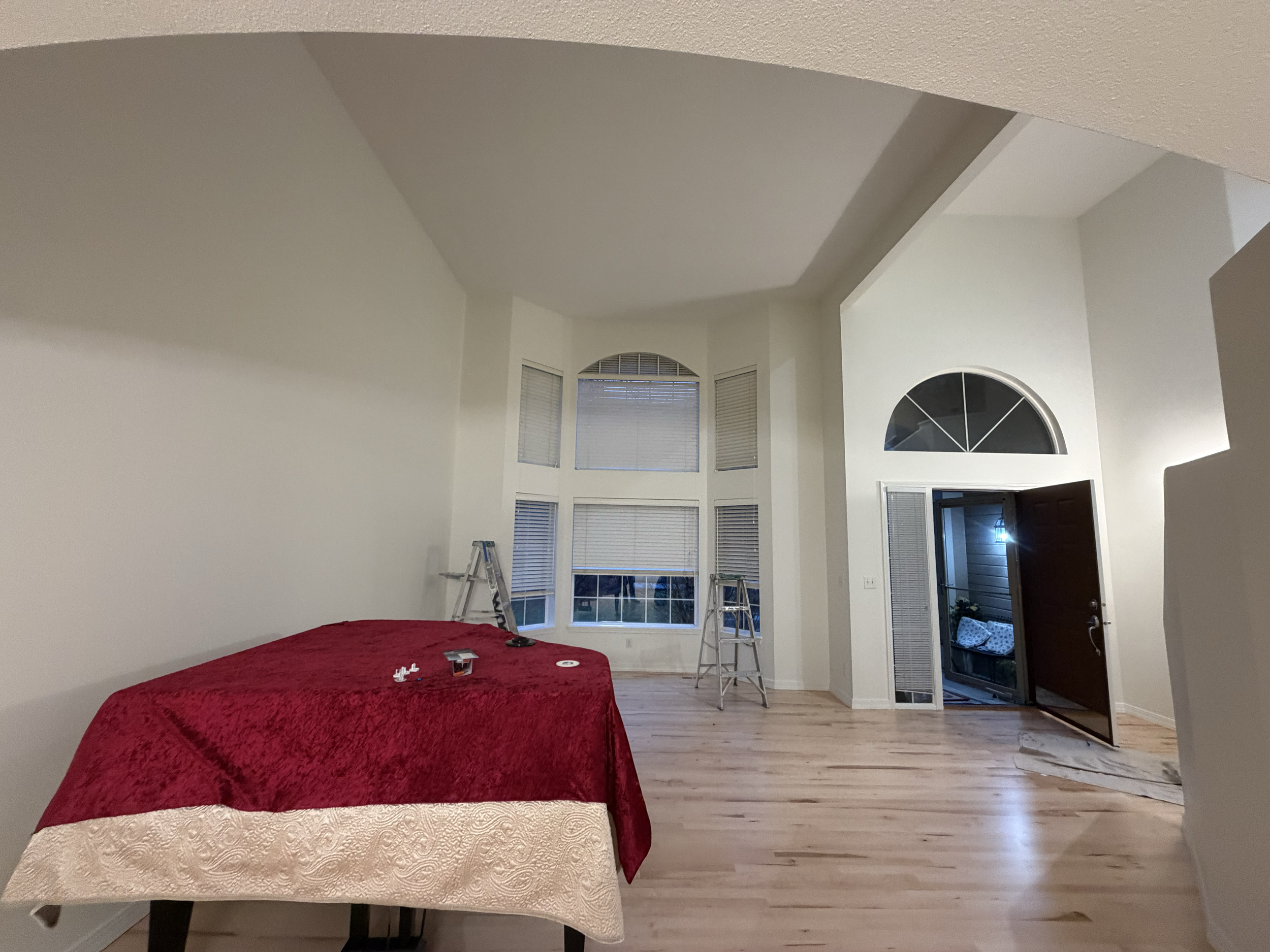 Interior view of a house with a high ceiling, large bay windows with blinds, hardwood flooring, an open doorway leading outside, and a table with a red tablecloth in the foreground. Luxury residential home painted in Kuna, Idaho.