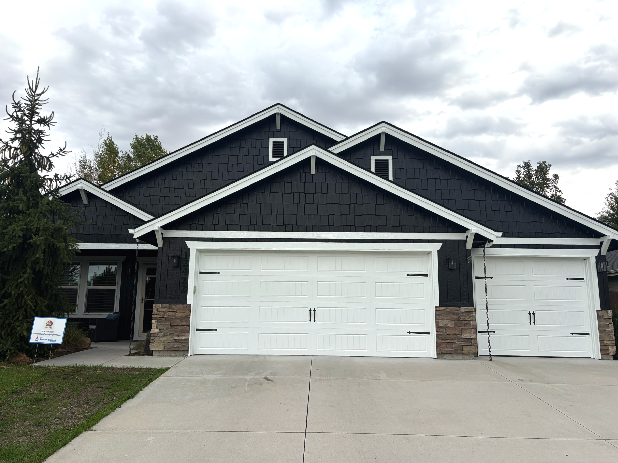 Front view of a modern house with black shingle siding, white trim, and a two-car garage with white doors. There are two vents on the gable ends of the roof, and a small tree on the left side of the house. The driveway and walkway are concrete, and the sky is cloudy.
