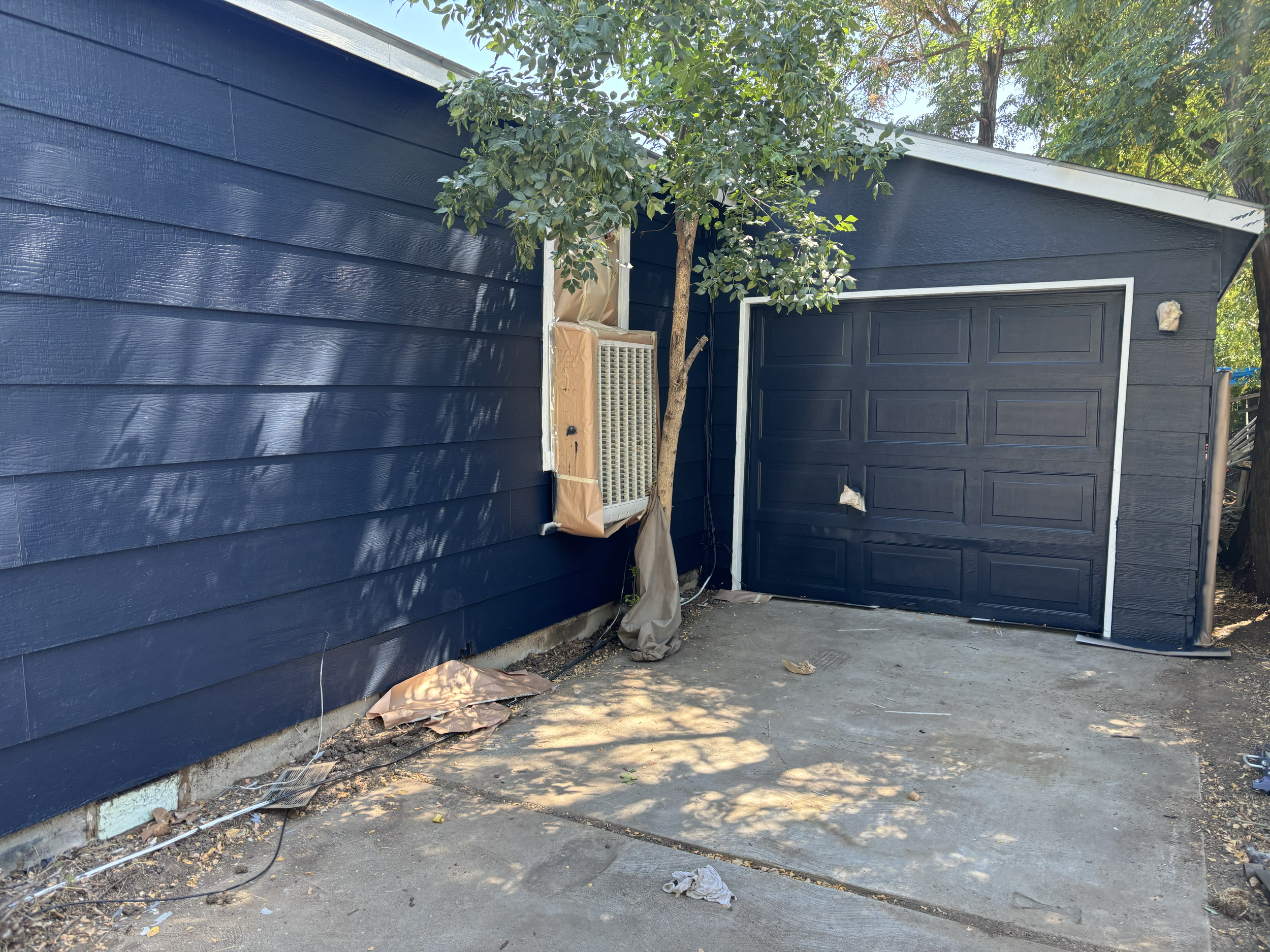 Blue house with horizontal siding, a tree growing close to the house, an air conditioning unit covered with paper, and a closed garage door. Debris and tools are scattered on the ground outside. Residential exterior painting in Boise, ID