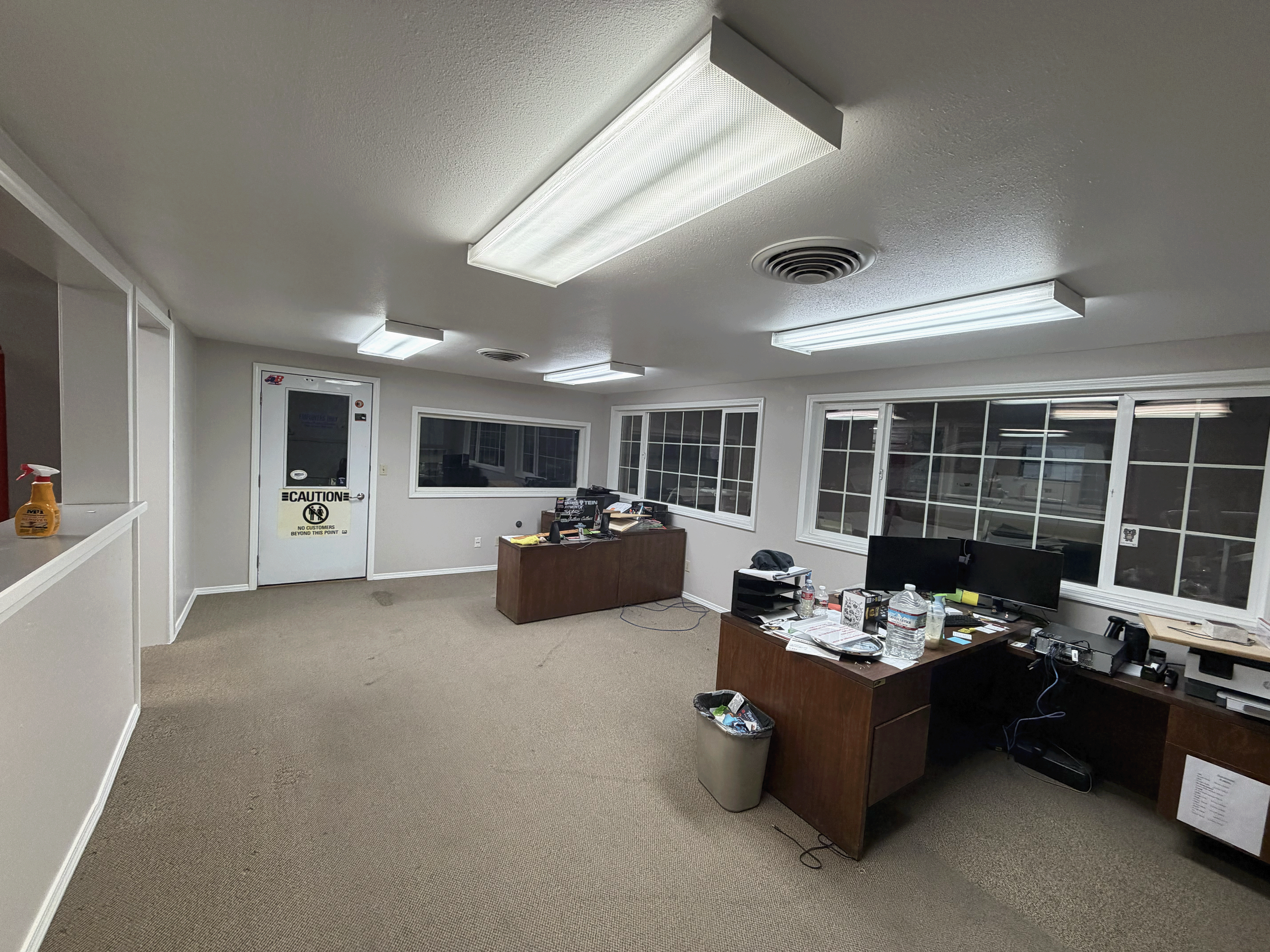 An office room with beige carpet, white walls, and ceiling. There are four rectangular fluorescent ceiling lights, windows along one wall, and a white door with a caution sign. The room has desks with computer monitors, water bottles, papers, and ele