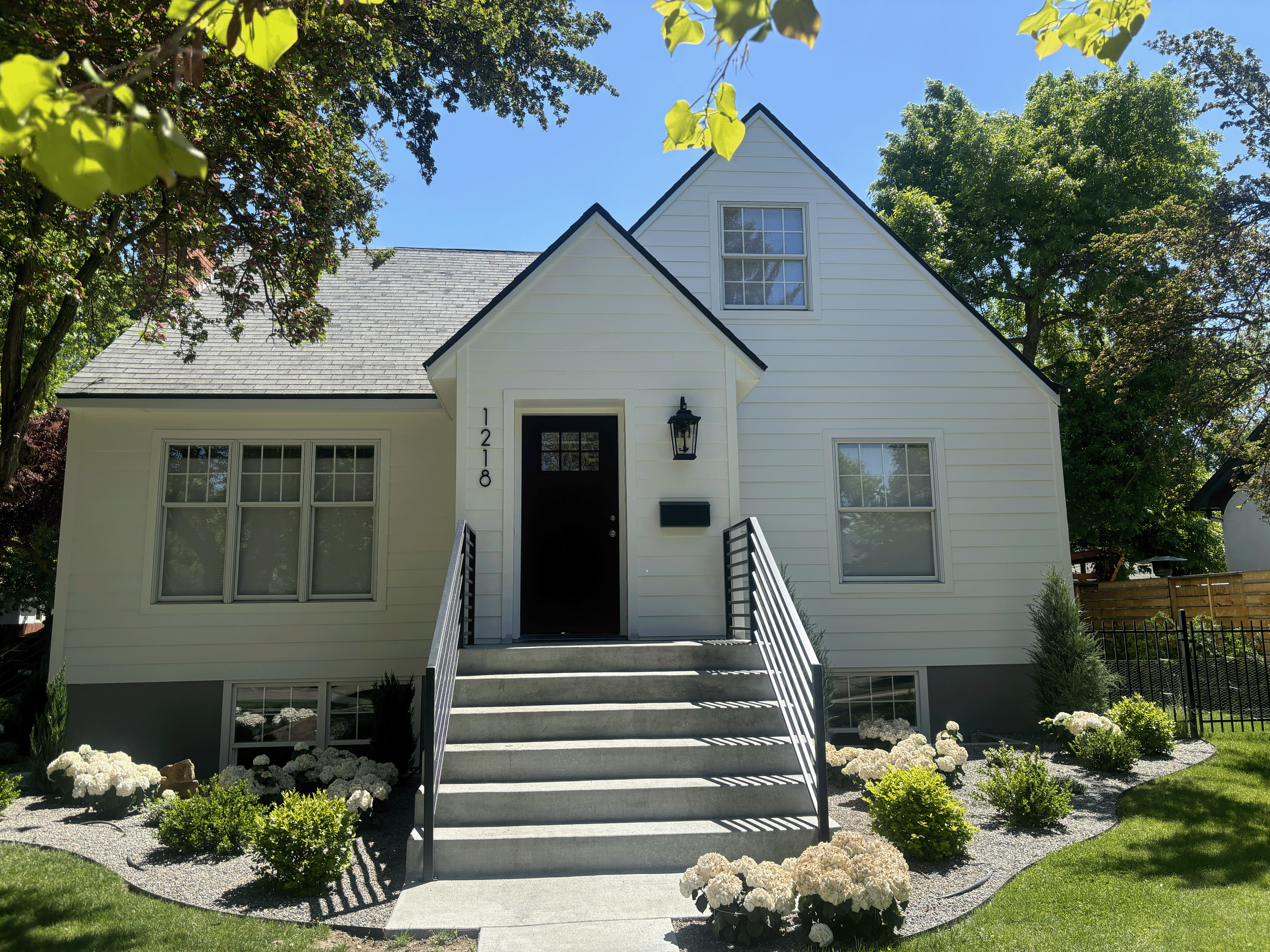 A exterior painted white house with black window frames and a black front door, surrounded by a lush garden with white flowers and green shrubbery, and tall trees with green leaves in the background under a bright blue sky.