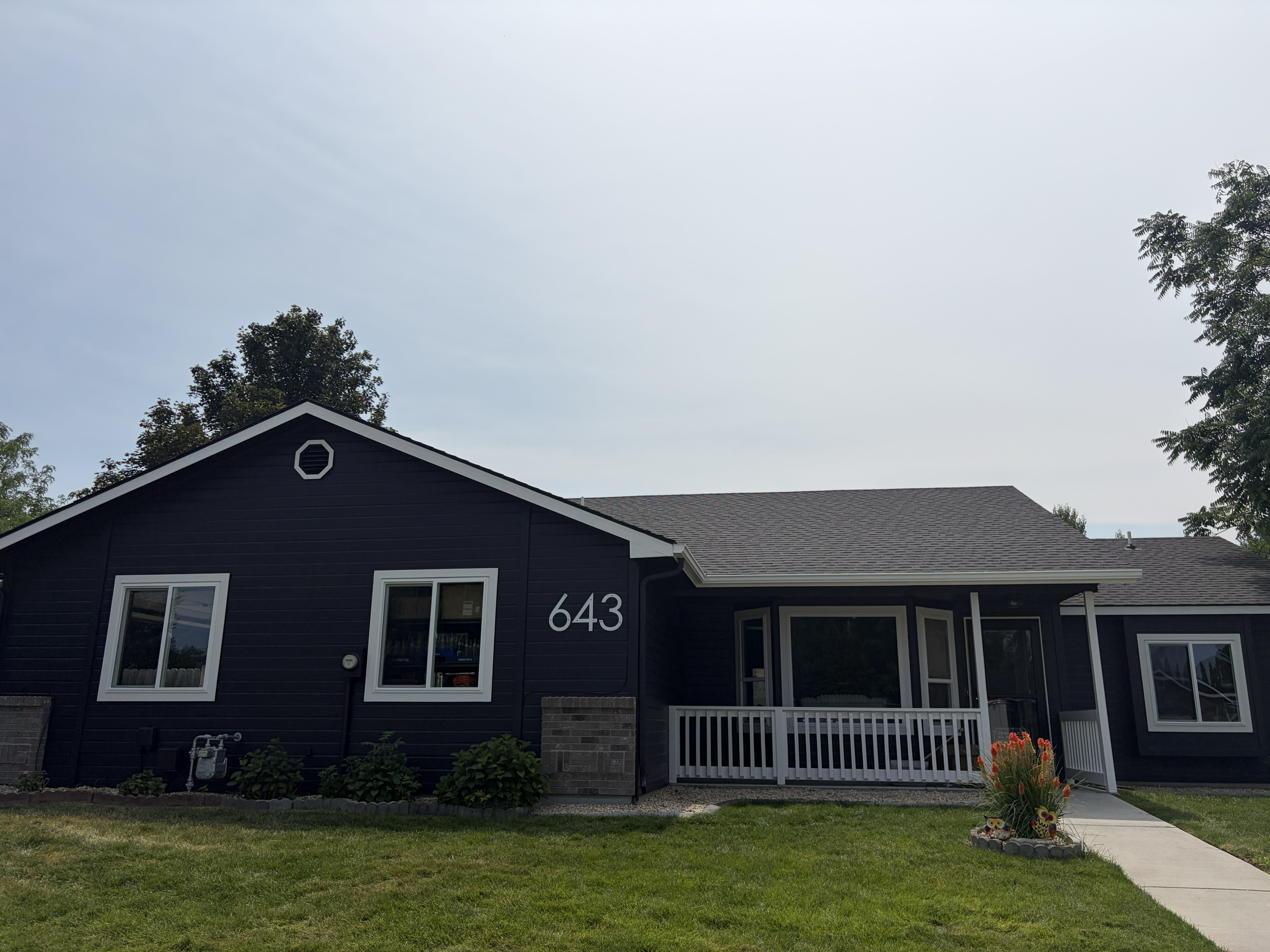 Front view of a dark blue house with white-framed windows, a small porch, and a yard with green grass and flowers, with trees and a cloudy sky in the background. Freshly painted in Boise, Idaho