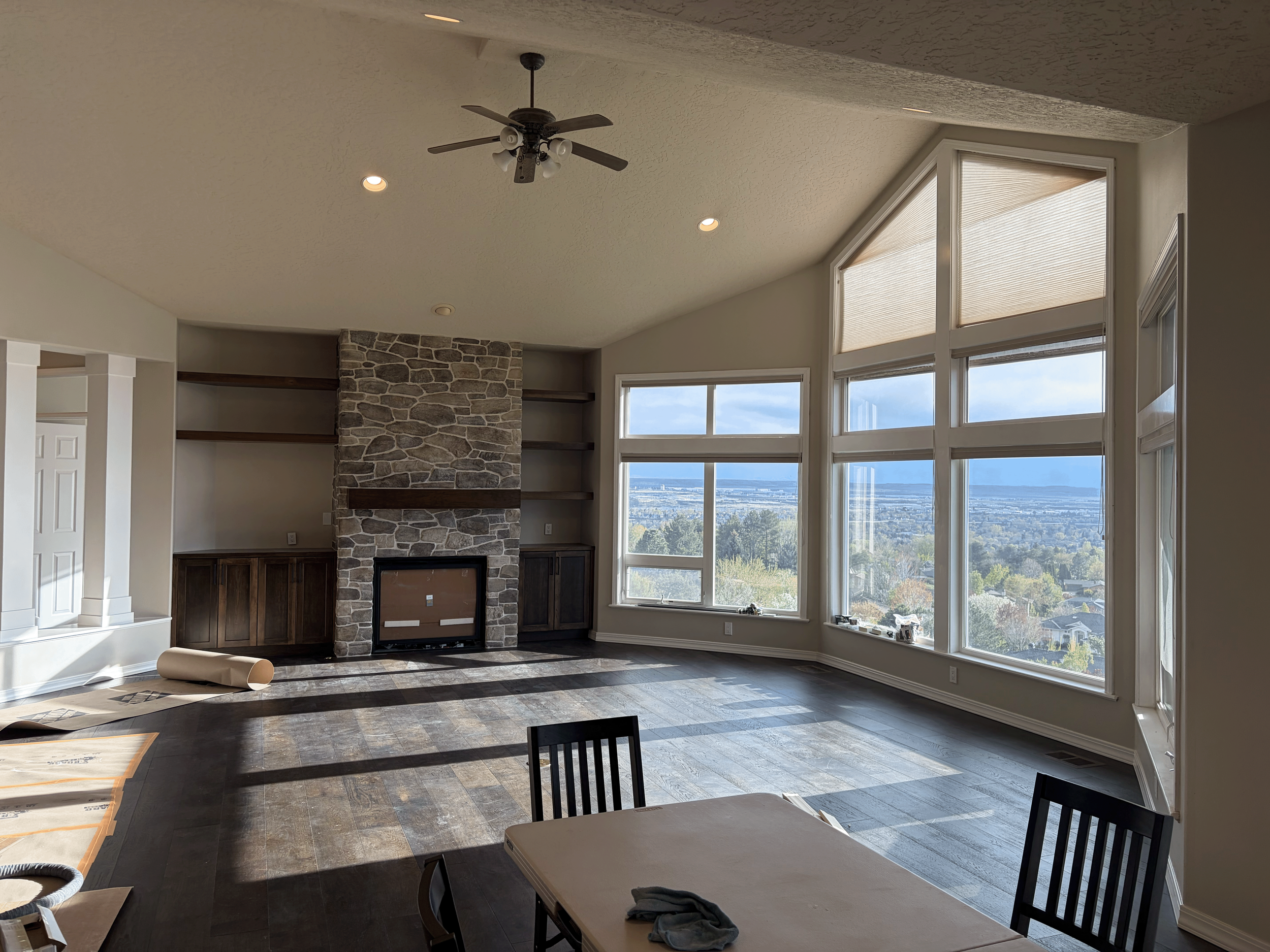In progress tan painted living room with large windows, stone fireplace with wooden mantel, built-in shelves, ceiling fan, and view of a landscape outside. Luxury home painting
