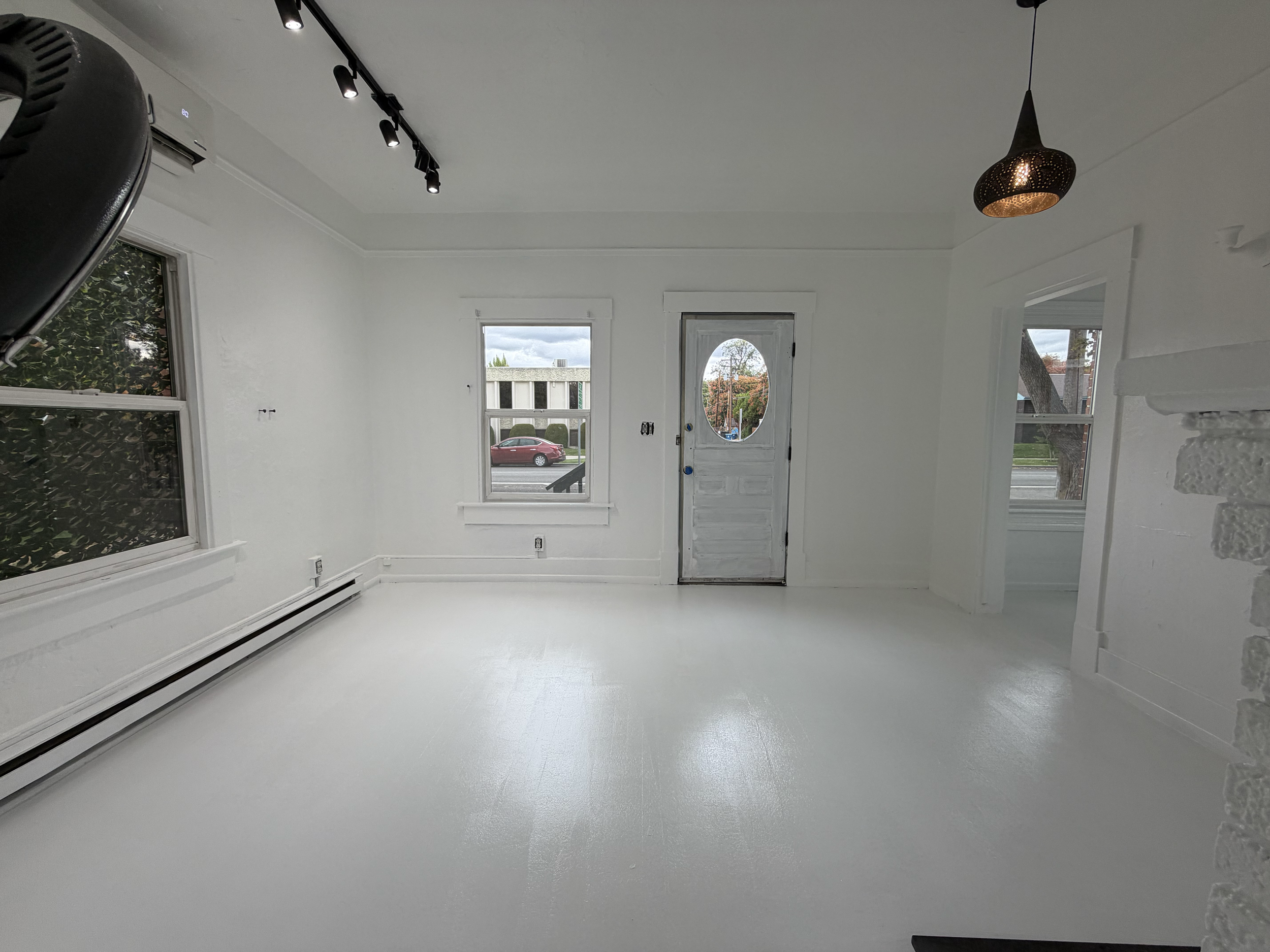 Completed interior painting in Caldwell, Idaho. Empty room with white walls, large windows, and a white door with a window. Ceiling-mounted track lighting and a hanging lantern light fixture. Outside view of street with trees and cars.