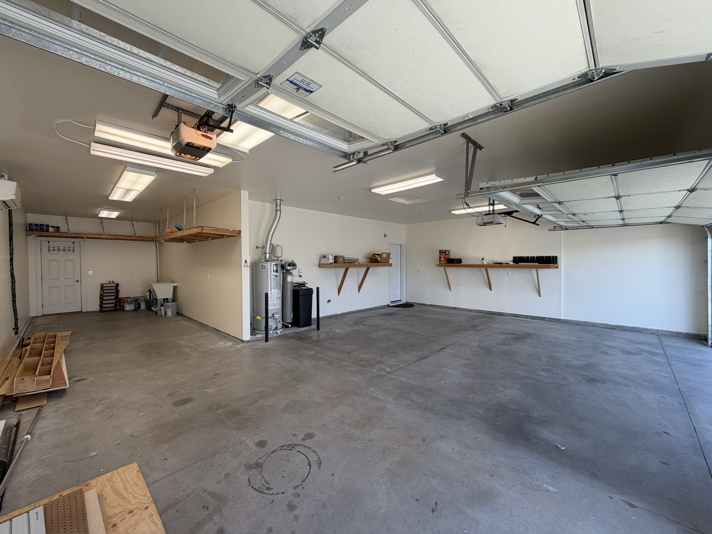 Empty garage with a partially open garage door, fluorescent ceiling lights, a water heater, shelves, and some boxes along the walls. Before epoxy flooring application in the Treasure Valley