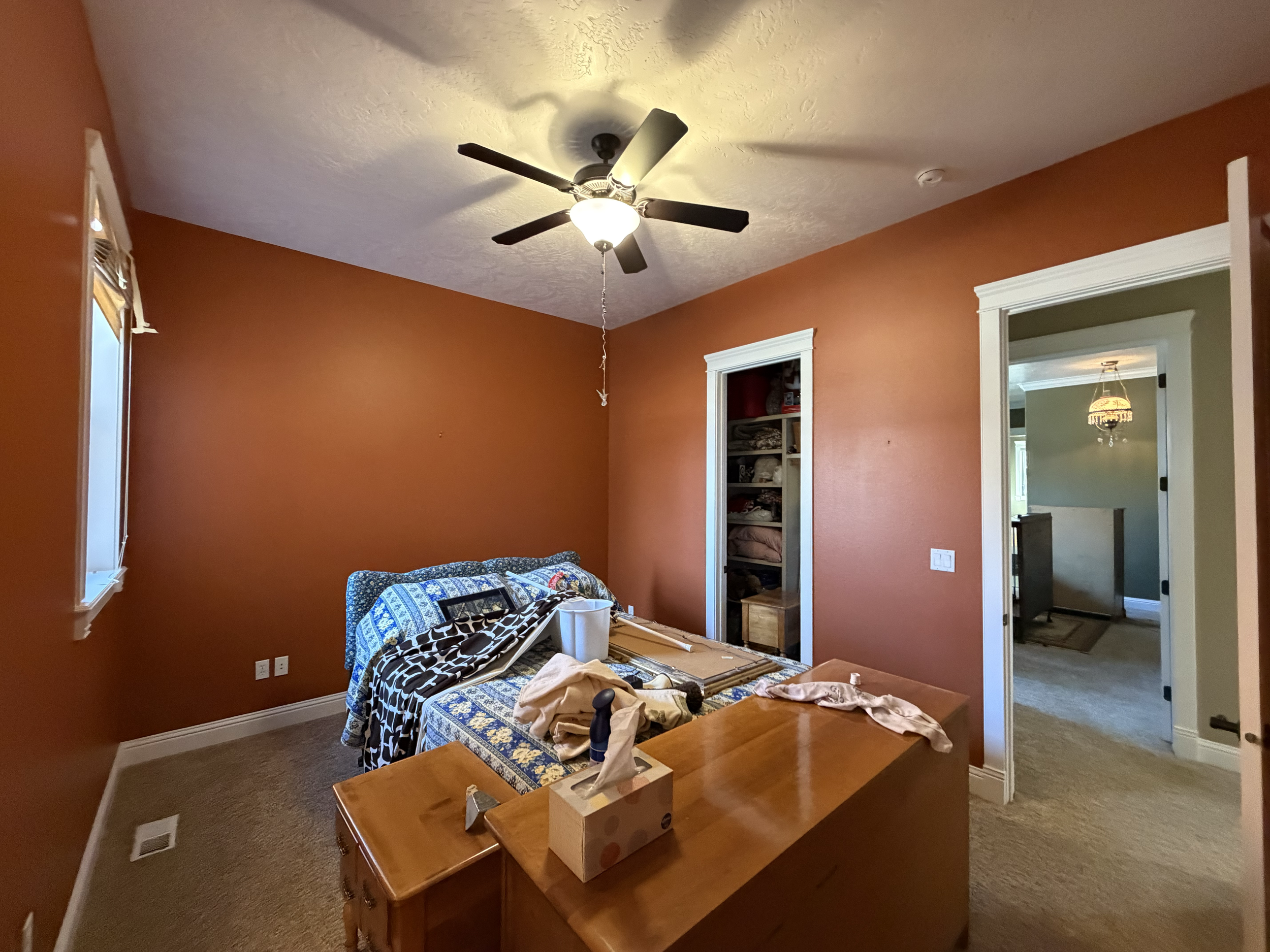 A bedroom with an orange wall, a ceiling fan with a light, a window on the left, a built-in shelf filled with folded towels, and a doorway leading to another room with a chandelier. The bed and dresser are cluttered with clothes and miscellaneous ite