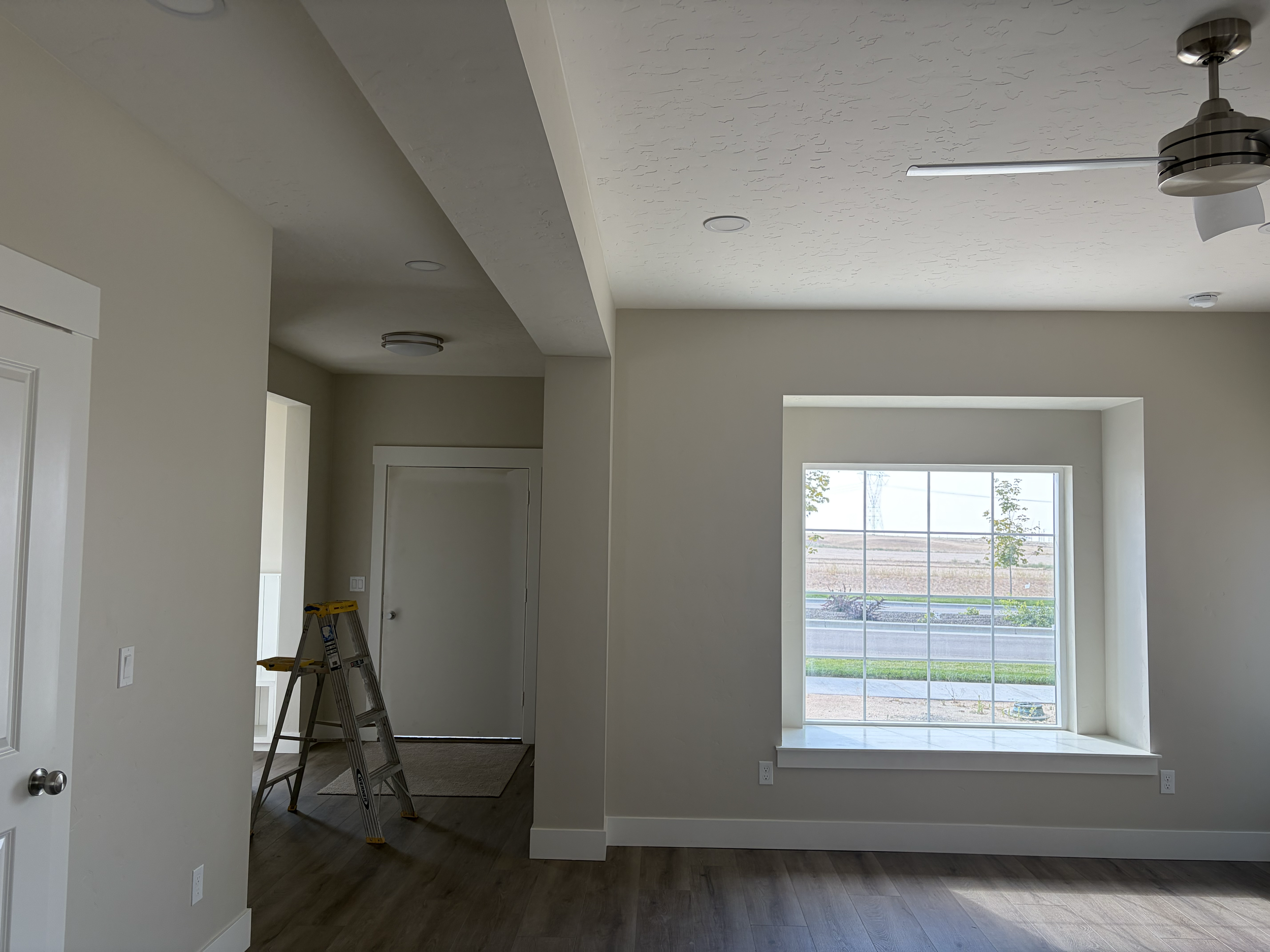 Interior of a new house with a large window, off-white walls, wooden flooring, and ceiling lights. A ladder is present near the doorway. Interior living room residential painting.