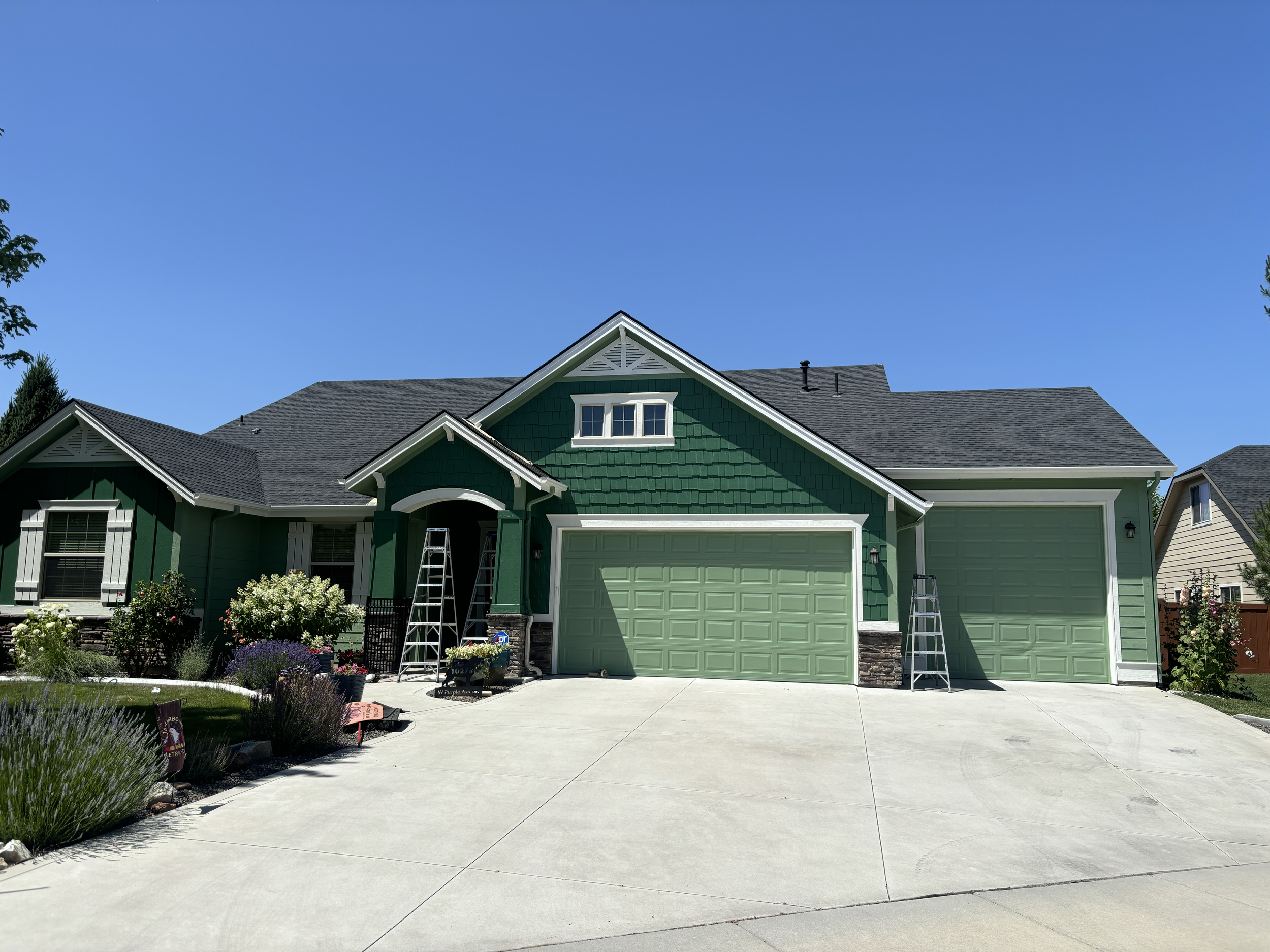 A two-story house with a green exterior, white trim, and a dark gray roof. The house has a garage, a front porch, and a garden with various plants and flowers. Two ladders are positioned on either side oft garage. Fresh, bright exterior house paint.