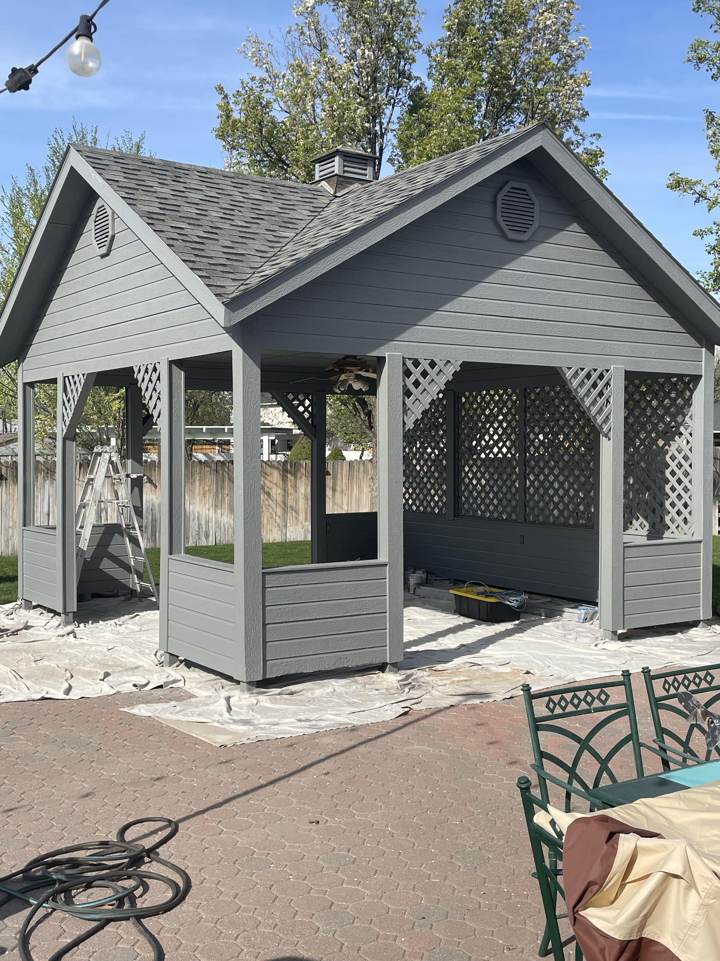 The image shows a backyard gazebo under construction, painted in gray, with a roof and lattice sides. There are tools and materials on the ground, a ladder leaning against the gazebo, and a string of outdoor lights hanging above. The area is covered 