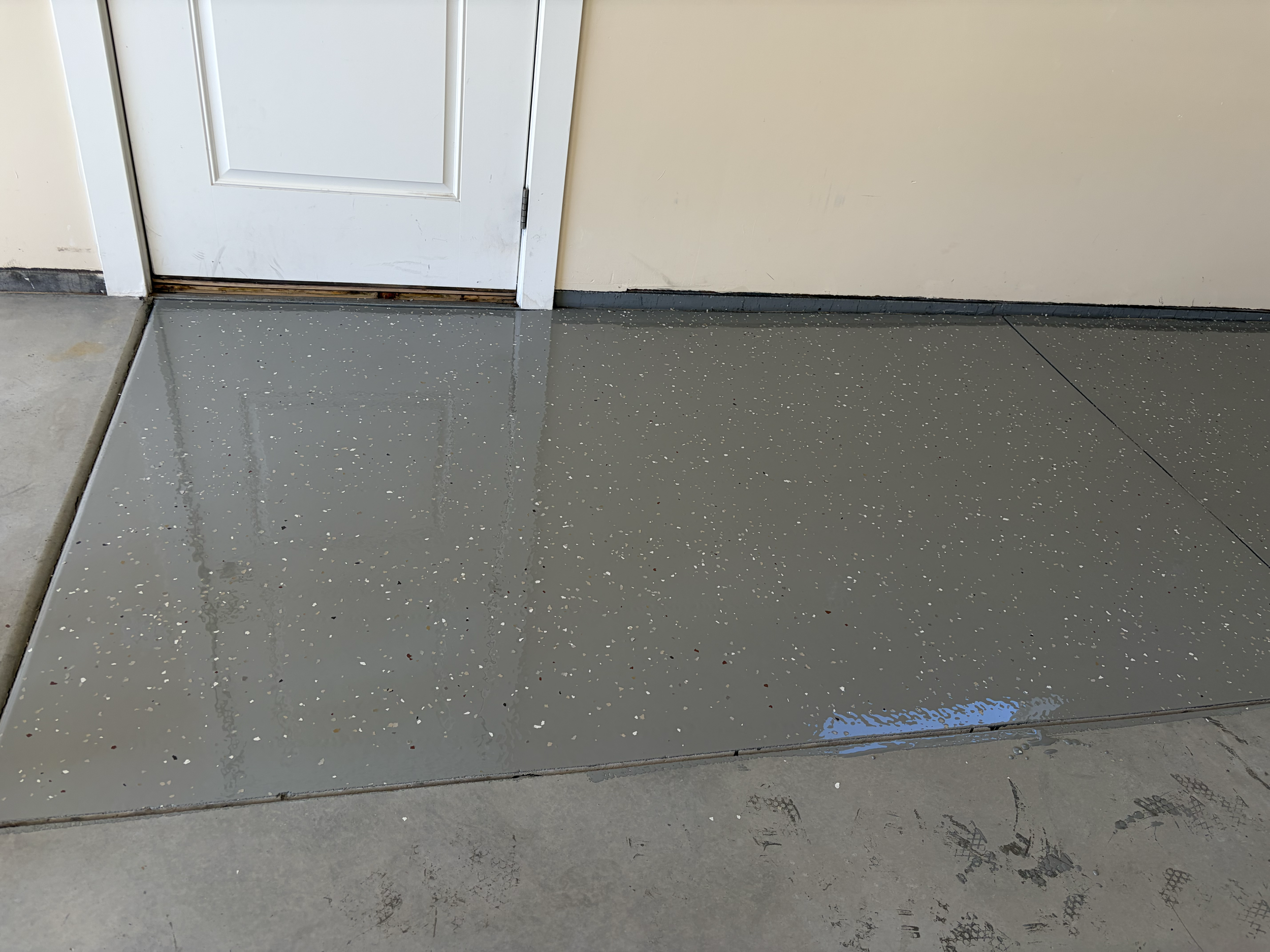 Close-up of a shiny gray epoxy speckled floor with a white door to a beige wall. The door has a small gap at the bottom showing a wooden threshold and carpeted flooring underneath. The epoxy is in progress of being cured. 