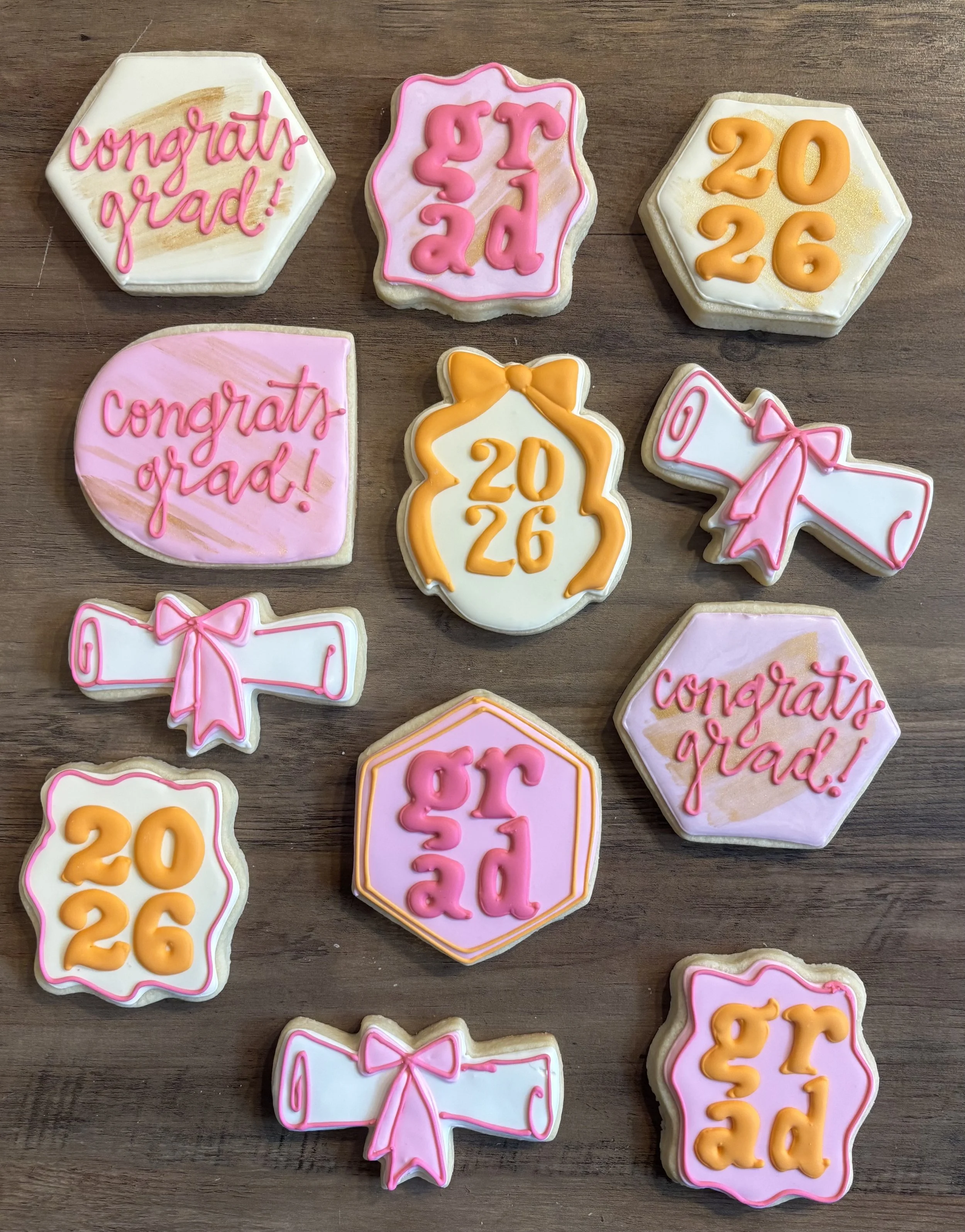 Rainbow-colored cookies with pink, orange, and yellow icing and message decorations in various shapes like hexagons and bows, celebrating graduate achievements with 'congrats grad!', '2026', '2024', and '2023' written on them.