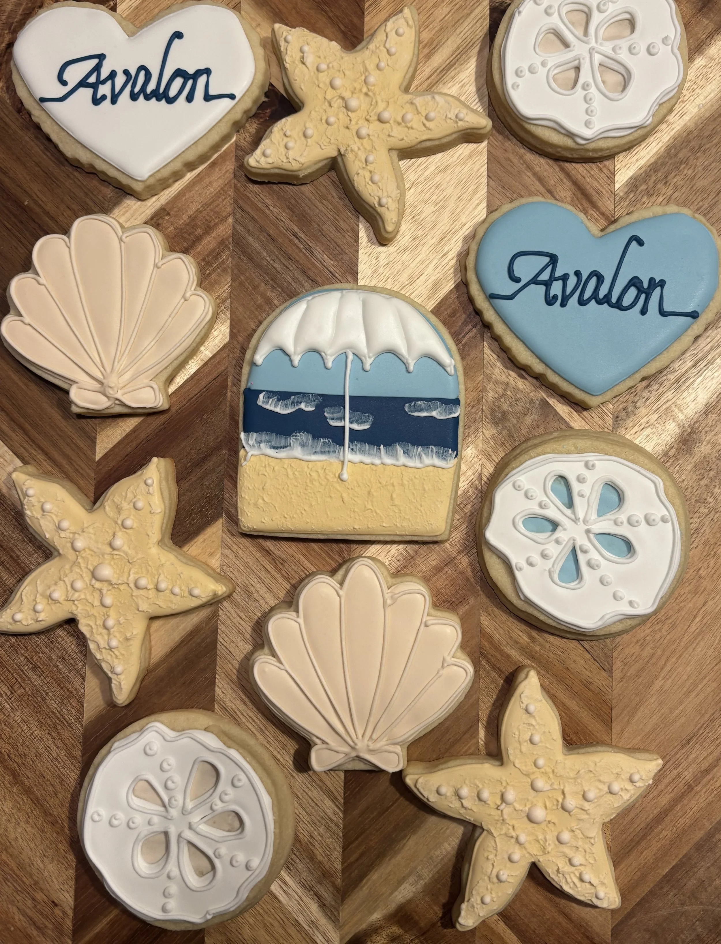 Collection of decorated cookies with beach and summer themes, featuring a starfish, seashells, a beach umbrella, and heart-shaped cookies with the word 'Avalon' written on them.