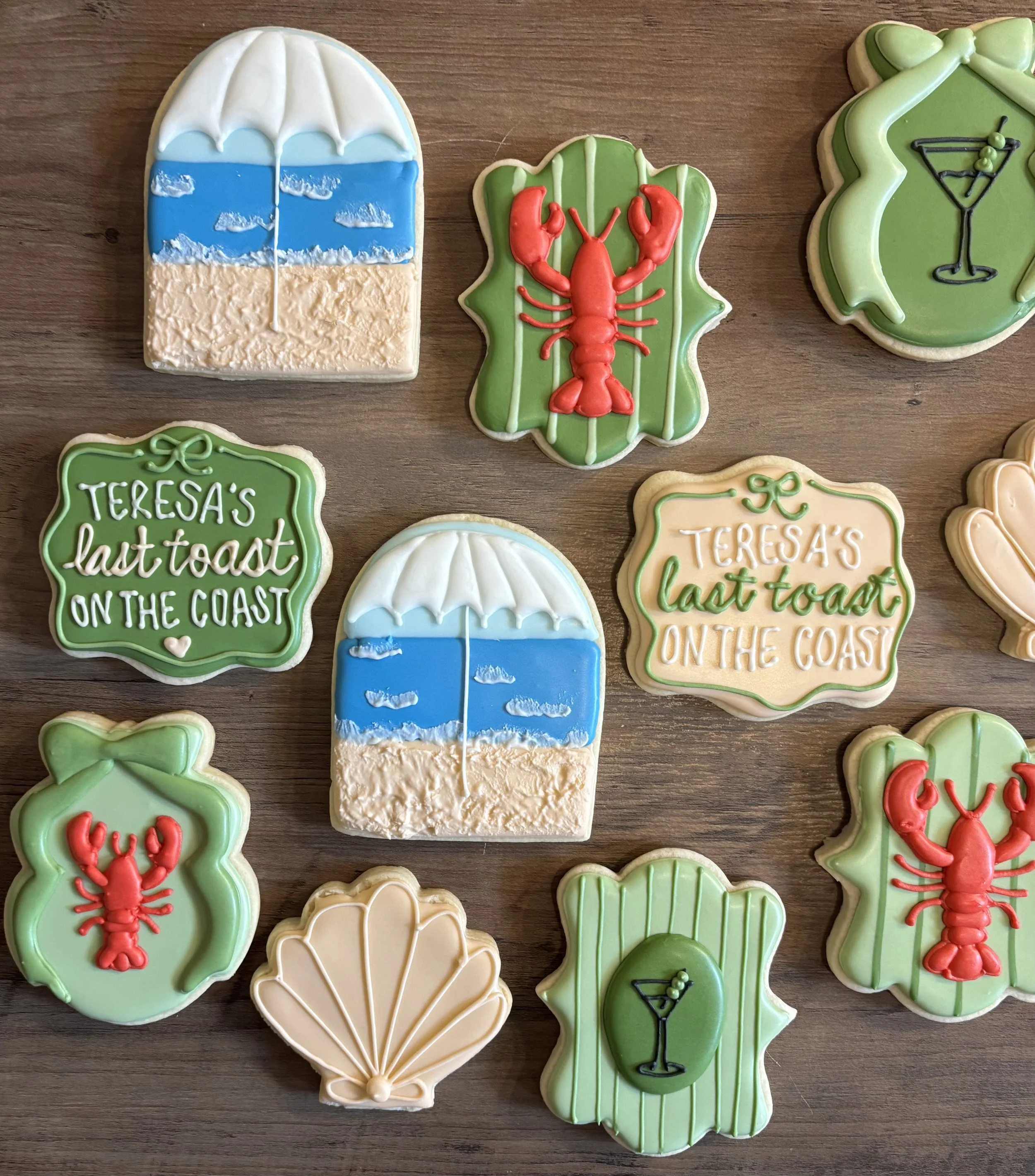 Decorative cookies on a wooden surface, including a beach scene with an umbrella, CGI lobster on a leaf, a cocktail glass on a lime green background, a shell, and a title with a green border and white lettering.