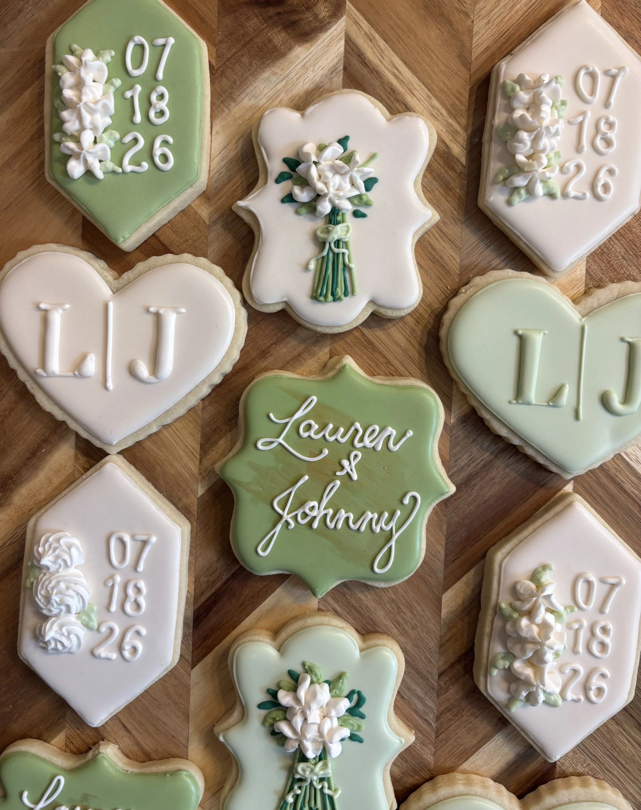 Decorated cookies with white icing and green accents, including date and name inscriptions and floral designs, arranged on a wooden surface.