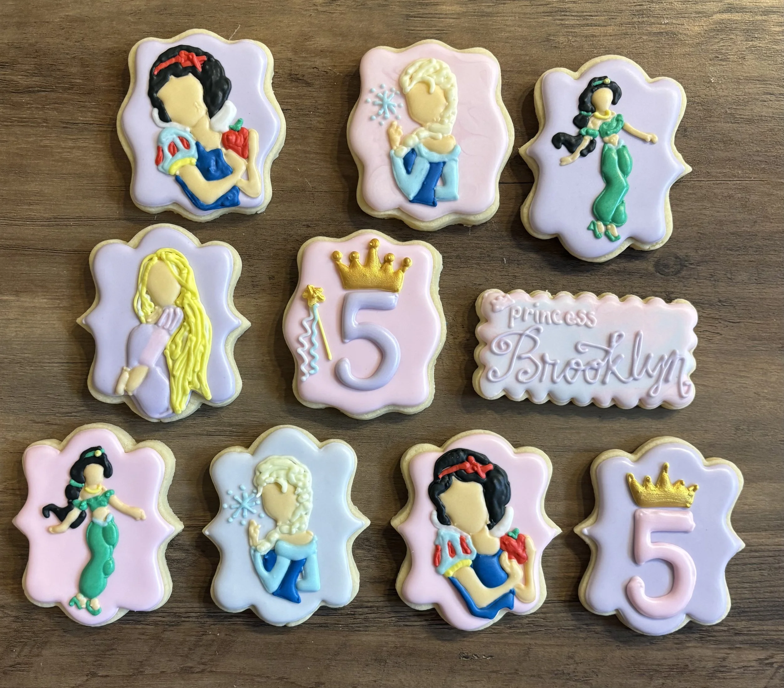 Collection of decorated cookies featuring princess characters, a crown, and the number 5 on a wooden surface.