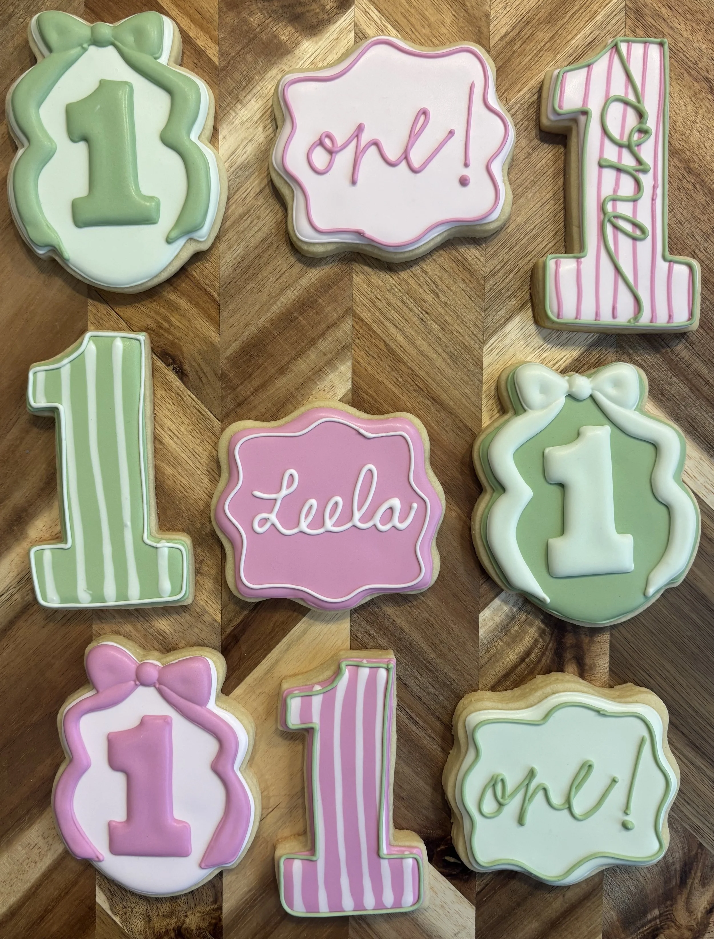 Decorated cookies celebrating a first birthday, with designs including the number one, a gift, and phrases like 'one!' and 'Lela', in pastel pink, green, and white icing.