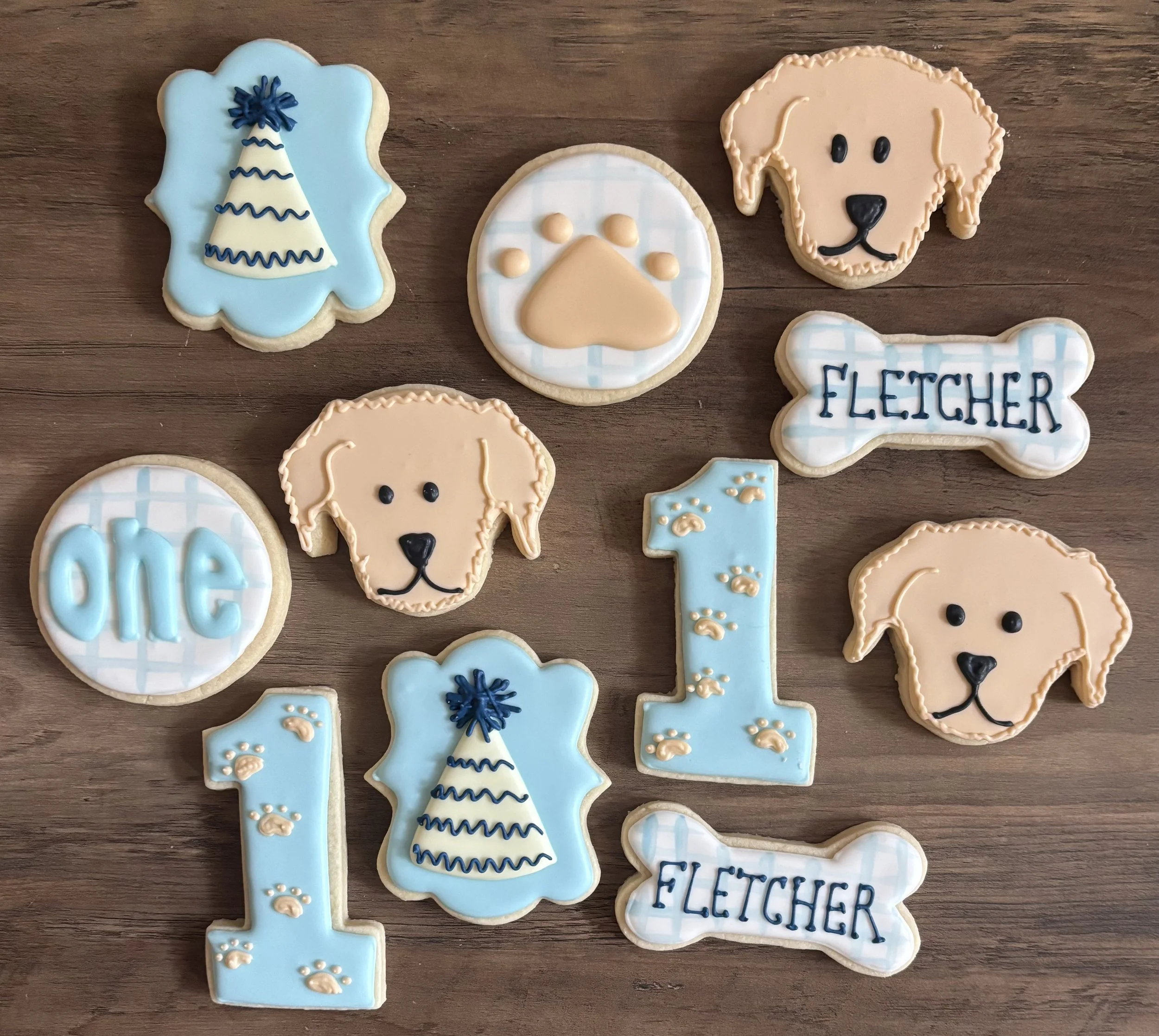 Cookies decorated with dog toys, a paw print, a dog bone, a dog face, and a sign that reads "Fletcher" in blue and beige colors.