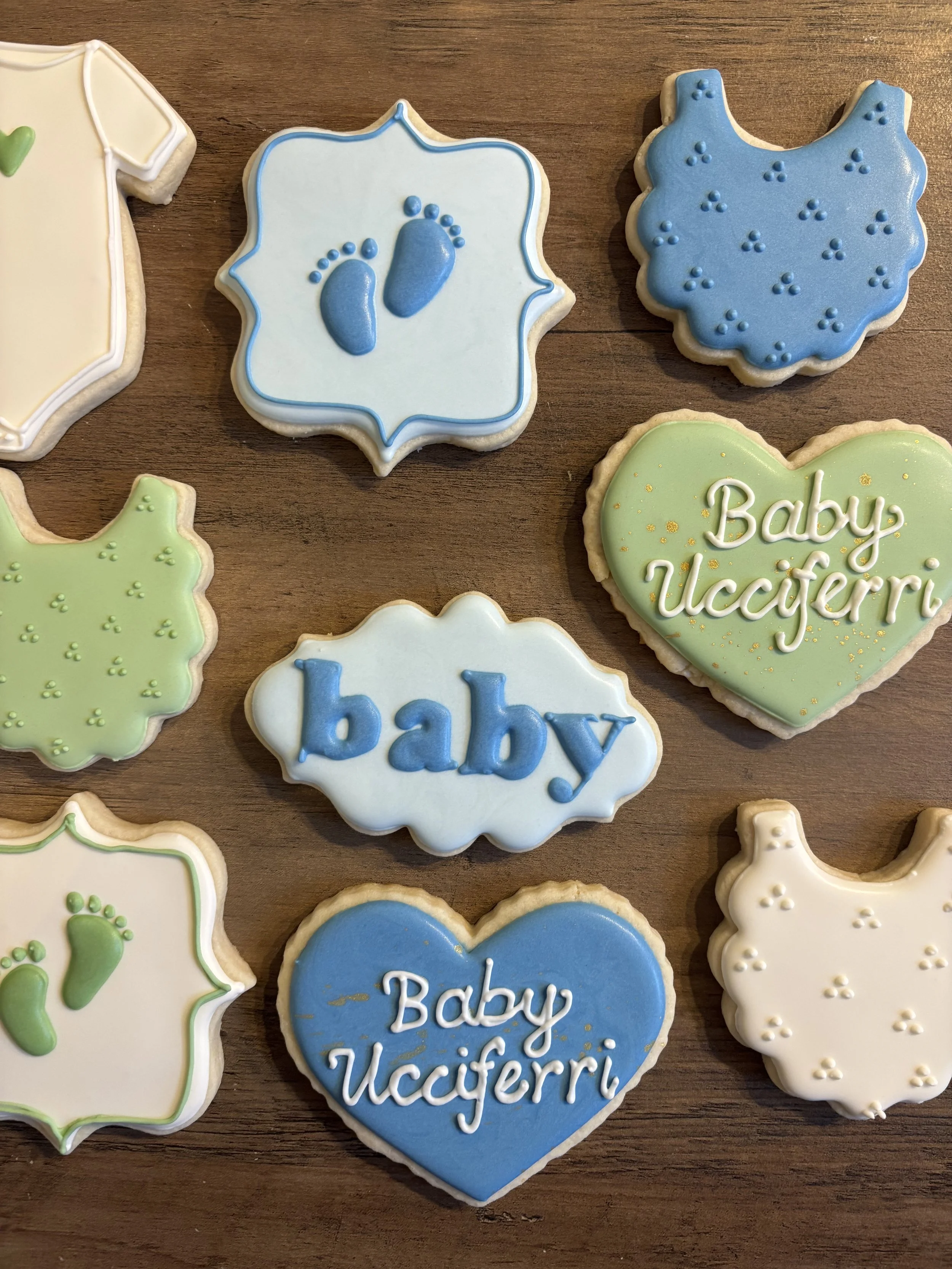 Decorative cookies on a wooden surface, including a white onesie with green baby footprints, a blue onesie with blue baby footprints, a green heart-shaped cookie with the words "Baby Ucciferri" written in white, and a white bone-shaped cookie with little white dots.