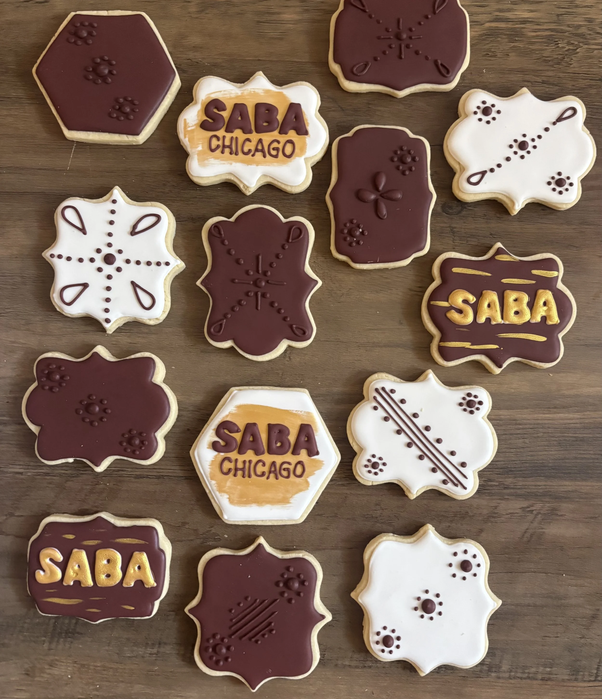 Assorted cookies on a wooden surface, decorated with icing and themed for SABAH CHICAGO, with designs of snowflakes, floral patterns, and the SABAH CHICAGO logo in yellow and brown.