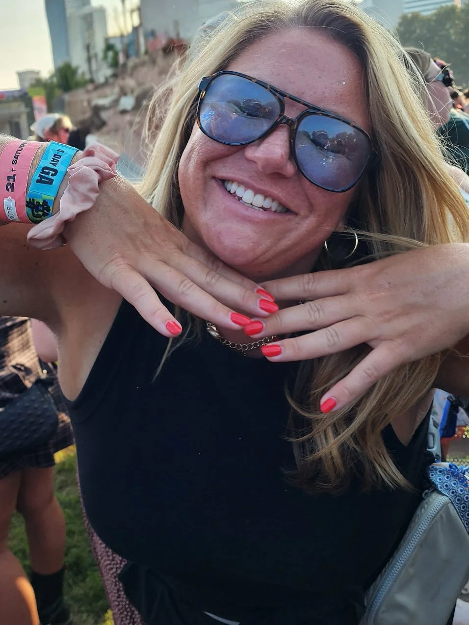 A smiling woman with blonde hair wearing sunglasses and red nail polish, holding a can of beer at an outdoor event.