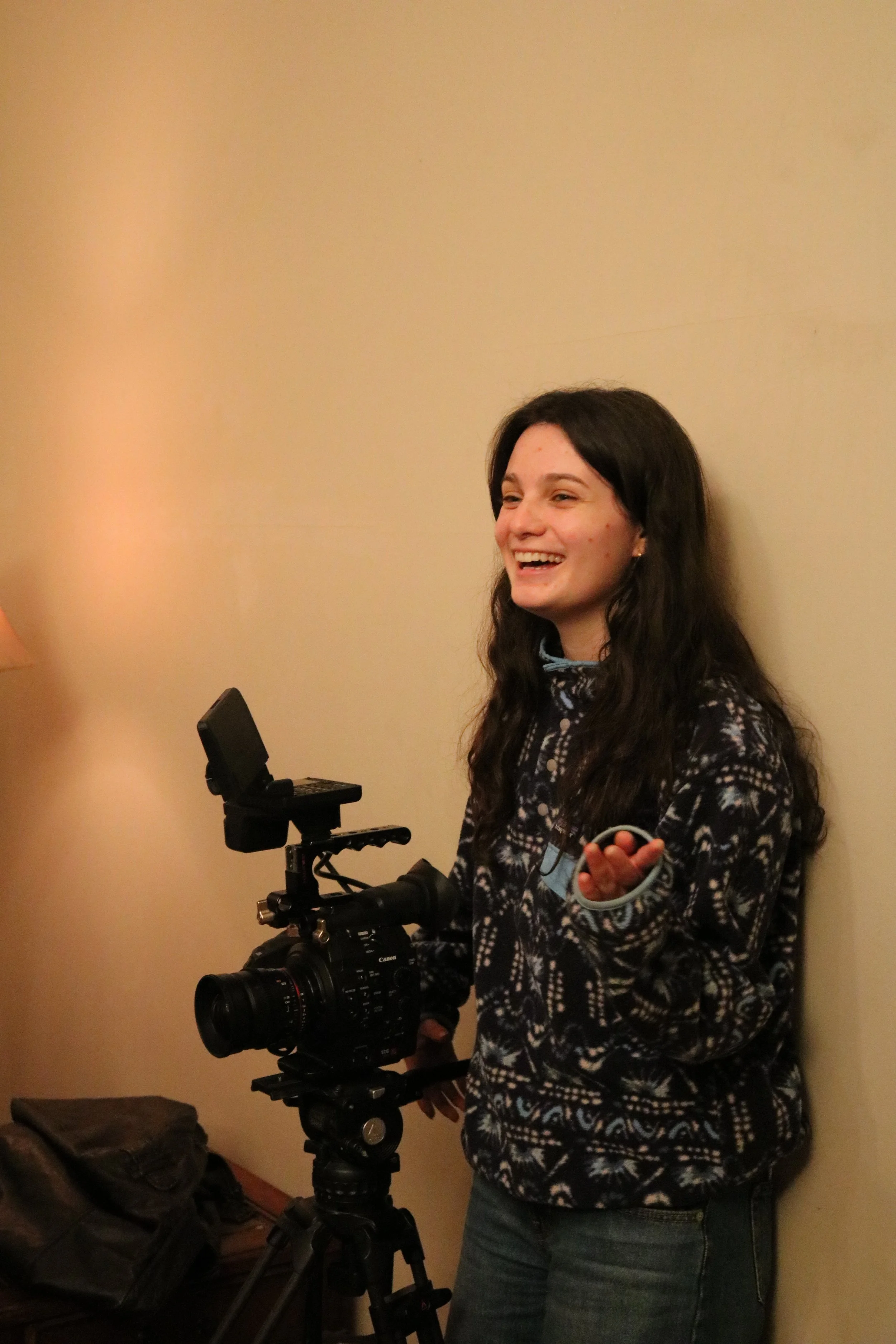 A young woman with dark brown hair, wearing a patterned sweater and blue jeans, smiling and standing next to a professional camera on a tripod, against a beige wall. There are some bags on the floor nearby.