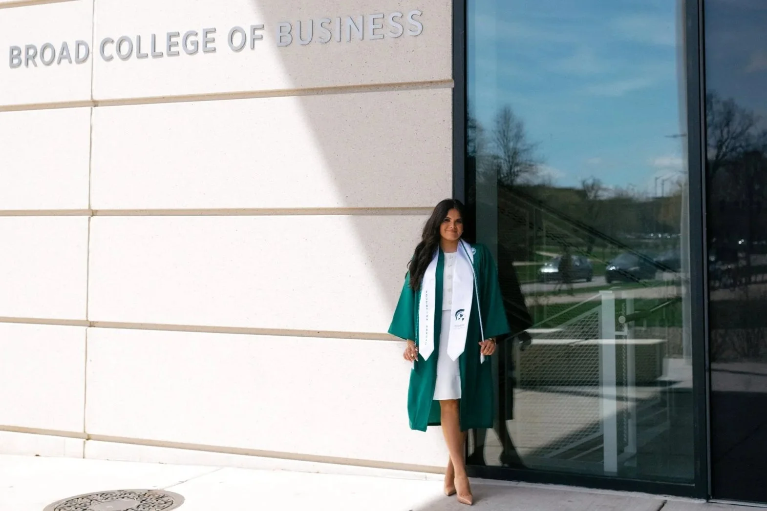 A woman in graduation attire standing outside the Broad College of Business, near a building with a sign and glass doors.