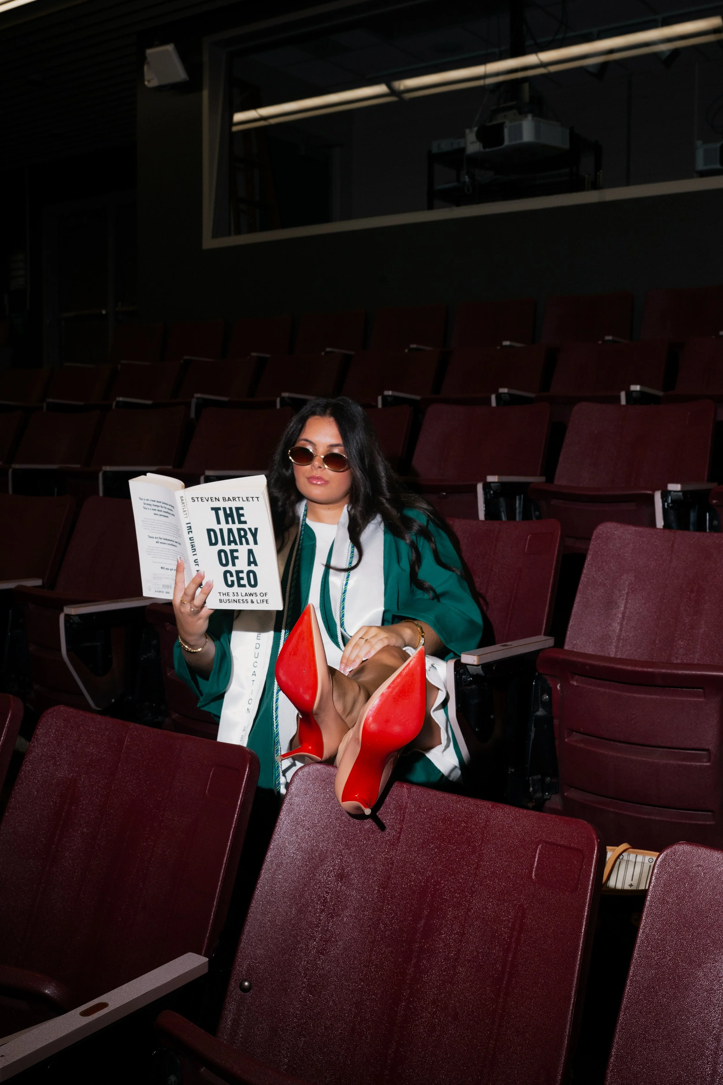 A woman with long dark hair, sunglasses, and wearing a green and white graduation gown is sitting in an auditorium with empty maroon chairs. She is reading a book titled 'The Diary of a CEO' and has red high-heeled shoes resting on a chair in front of her.