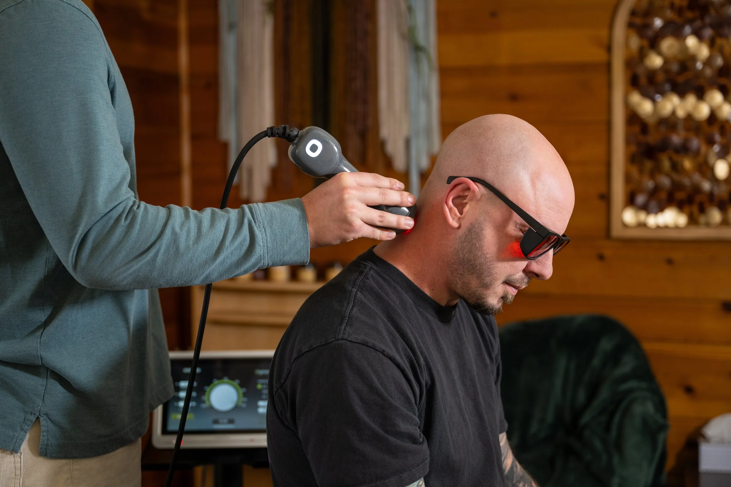 Person using a handheld laser therapy device on a bald man's neck in a log cabin