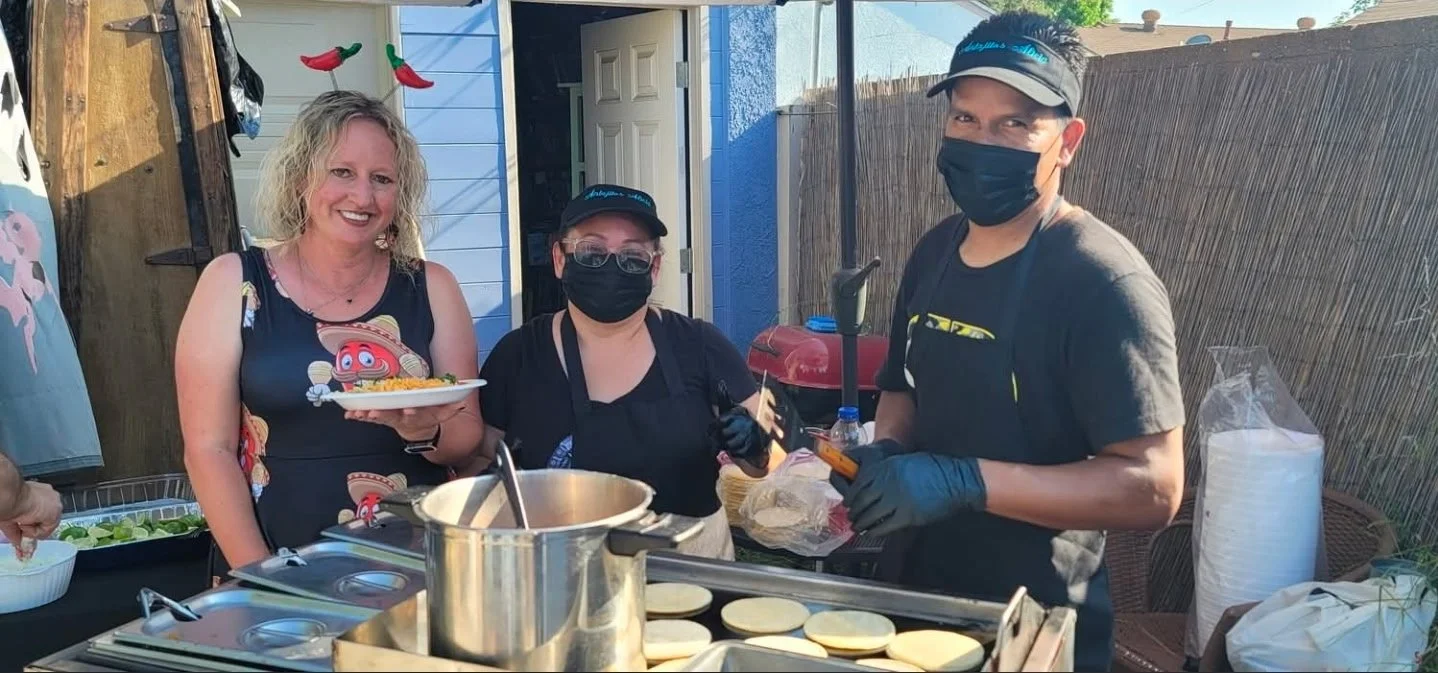 Three people in a backyard kitchen preparing and serving food, two wearing black masks and gloves, and one smiling woman holding a plate of food.