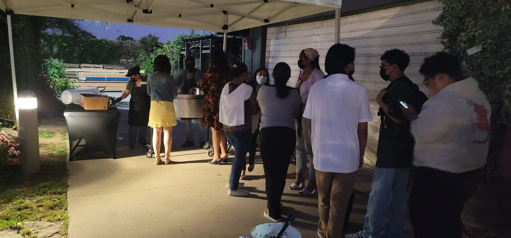 People standing in line at an outdoor food stand at dusk, with some wearing masks.