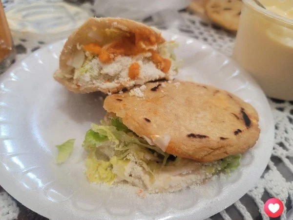 Half of a stuffed bread roll filled with vegetables and spices, and a pancake with some lettuce and sauce, on a paper plate with a side of creamy yellow drink in a cup.