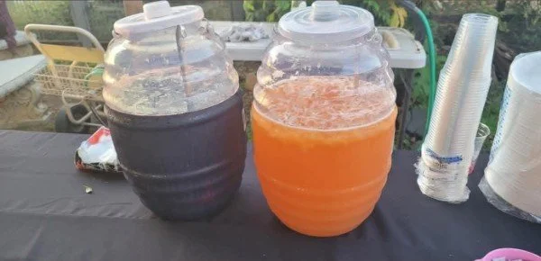 Two large jars filled with colorful beverages on a black table, with plastic cups stacked nearby.