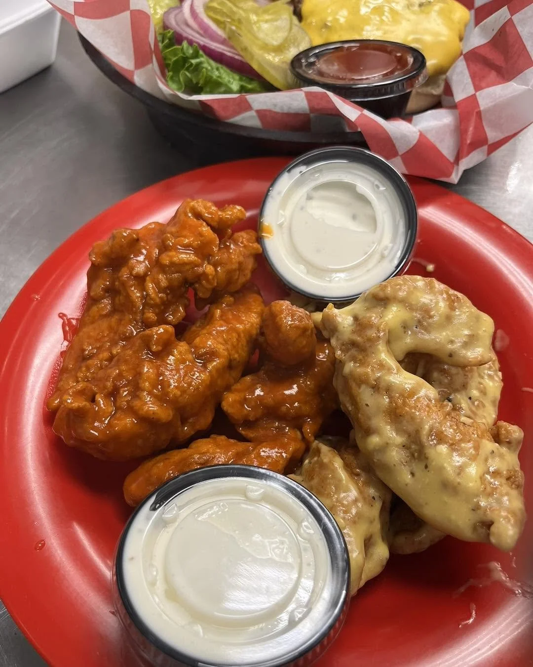A red plate withboneless chicken wings, some covered in spicy orange sauce and others in creamy cheese sauce. There are two small cups of ranch dressing on the plate. In the background, a basket holds a salad with lettuce, sliced onions, pickles, and