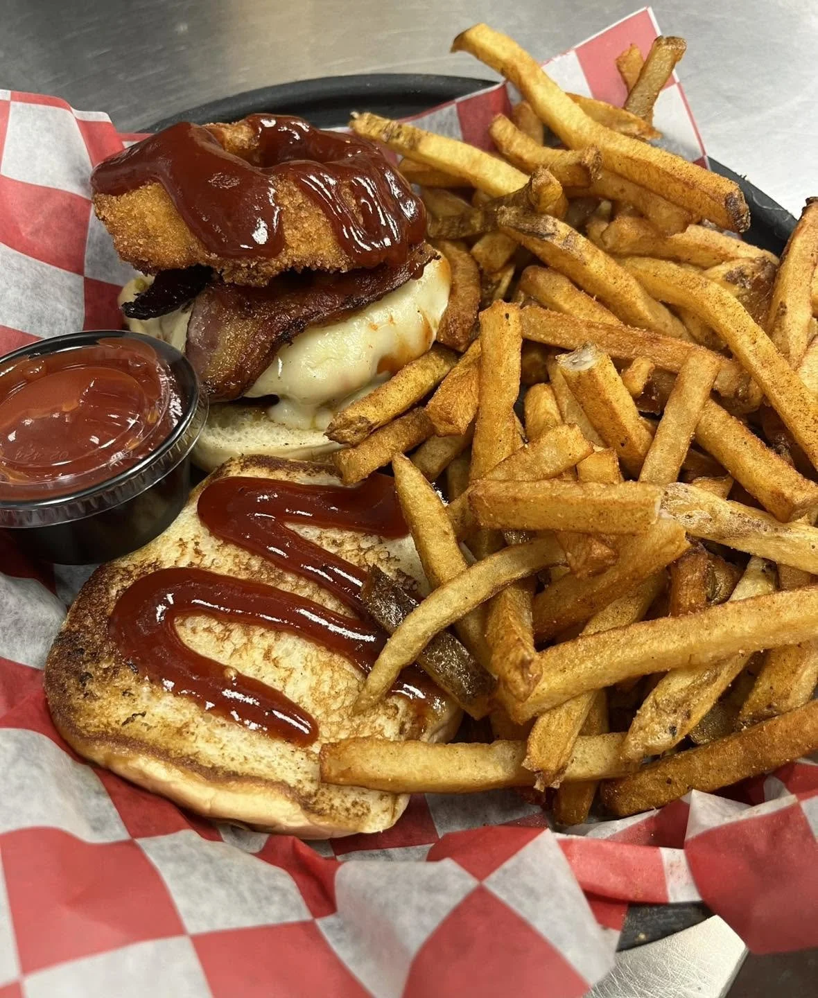 A basket of French fries, a sandwich with cheese, bacon, and ketchup, a fried chicken fillet with barbecue sauce on top, a toasted bread with ketchup, and a small container of ketchup.