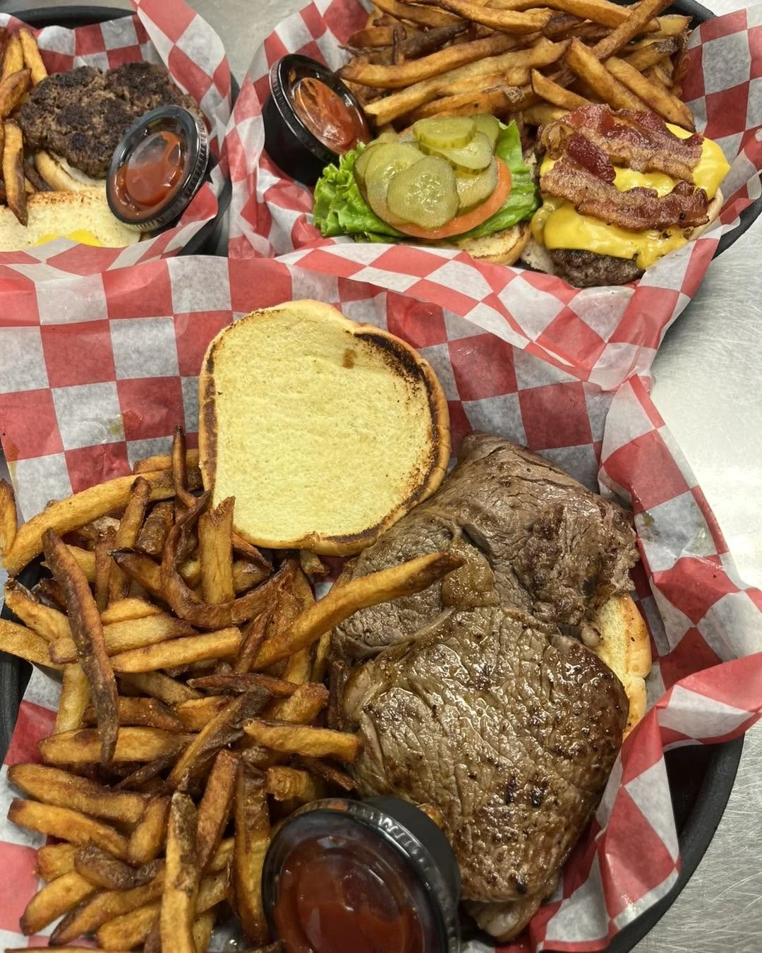 A tray with French fries, a burger with pickles, lettuce, tomato, bacon, cheese, and a beef patty, and a grilled steak, served with small containers of ketchup and barbecue sauce.