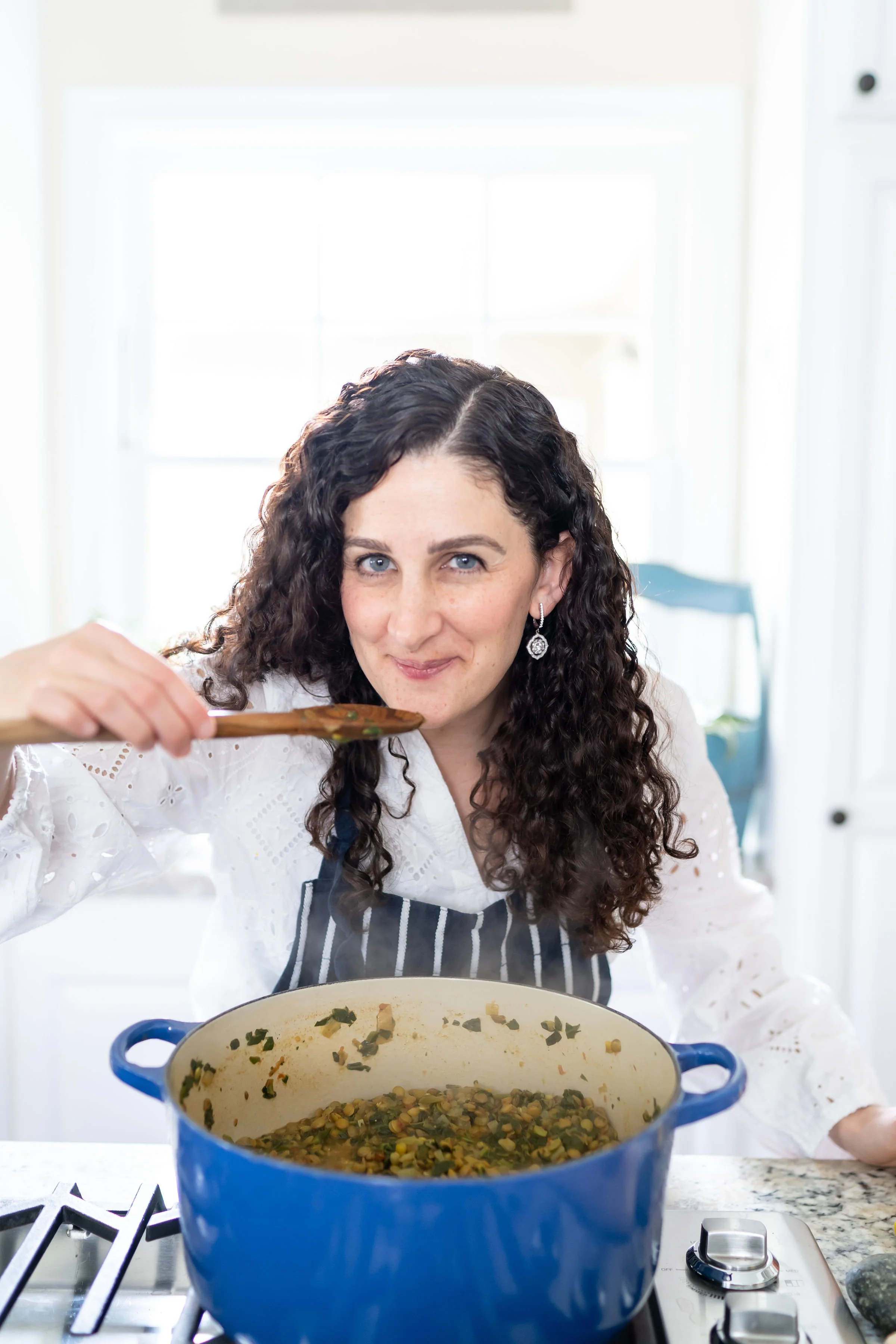 Woman with curly hair, wearing earrings and a white blouse, stirring a dish in a large blue pot on a stove in a bright kitchen.