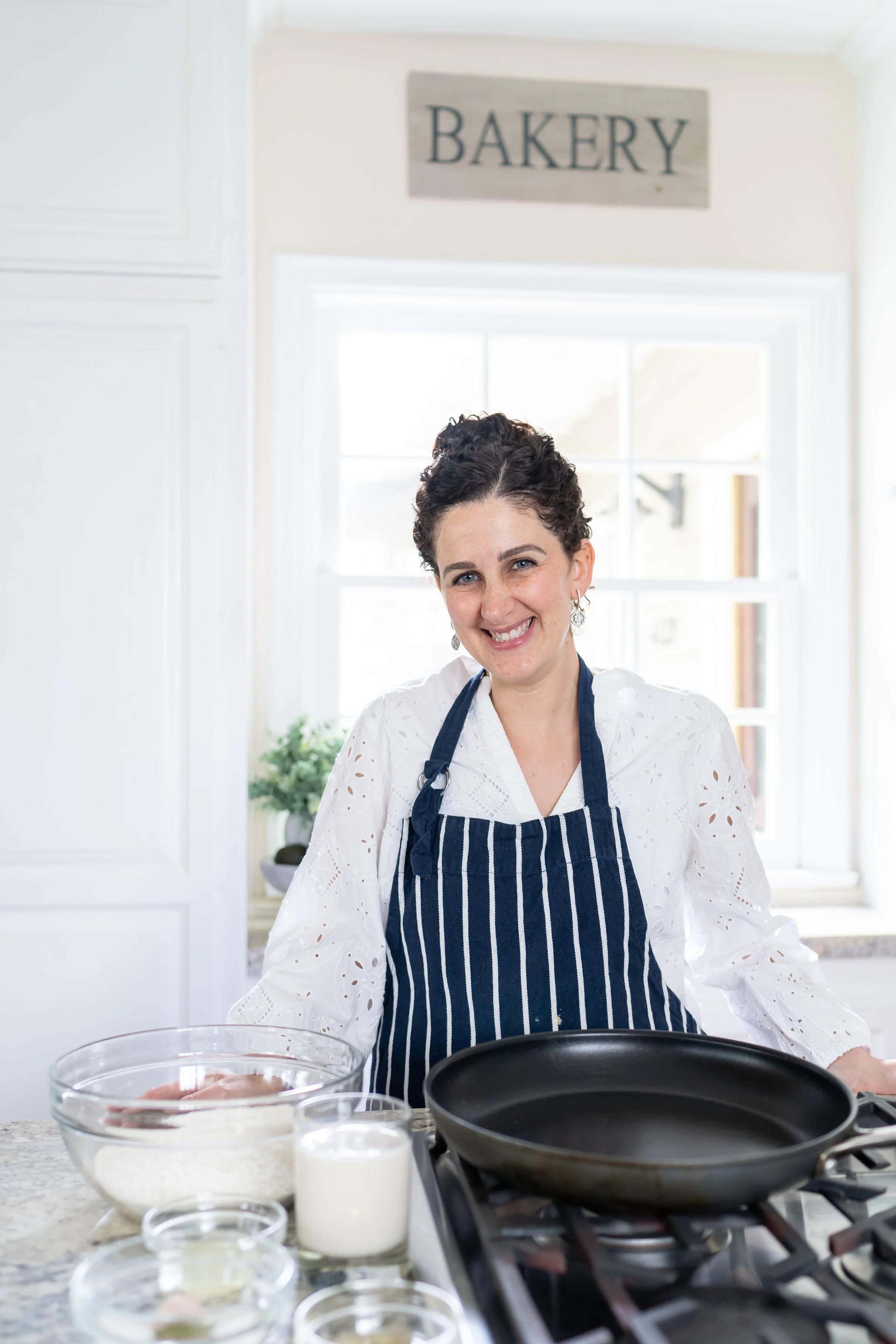 A woman with short curly hair smiling while cooking in a bright kitchen, wearing a white blouse with eyelet details and a dark blue striped apron, with baking ingredients and a skillet on the counter.