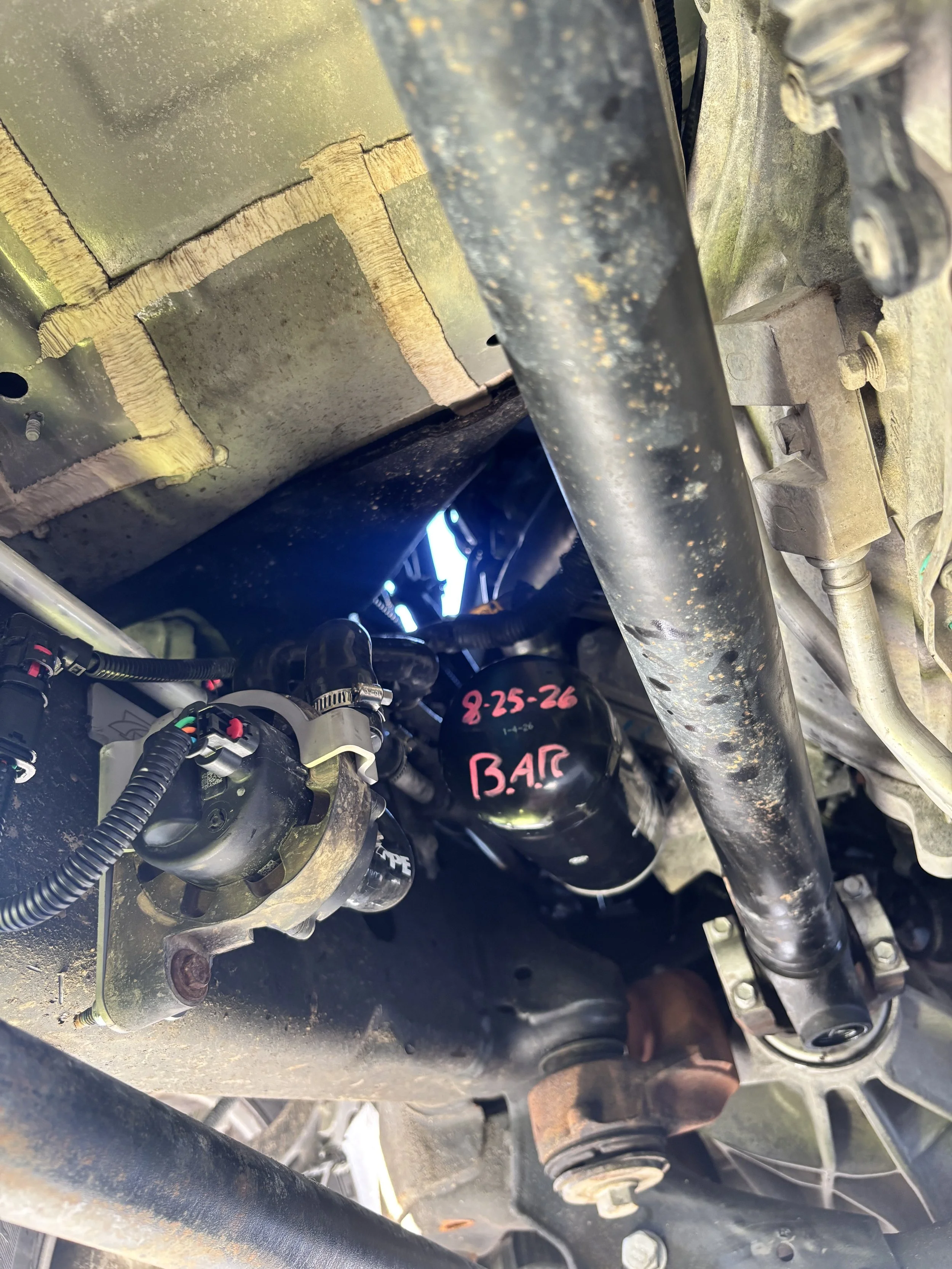 Underneath view of a vehicle's engine area showing parts including a black metal bar, hoses, and a component with writing '8-25-26' and 'B.A.P' in pink on a black surface.