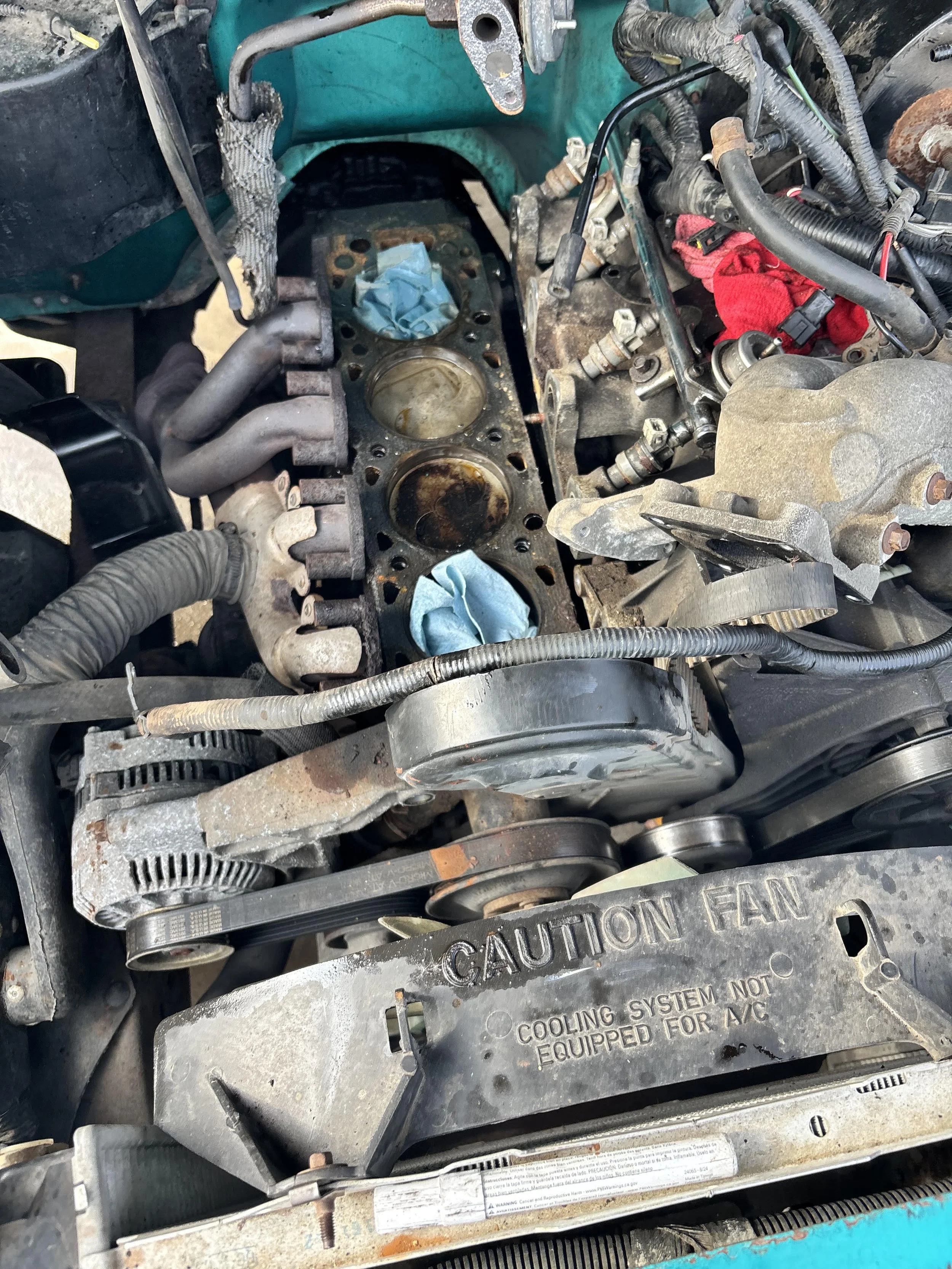 View of a car engine with the cylinder head removed, exposing the pistons and engine components, with some rust and dirt visible, and a warning label on the metal part.