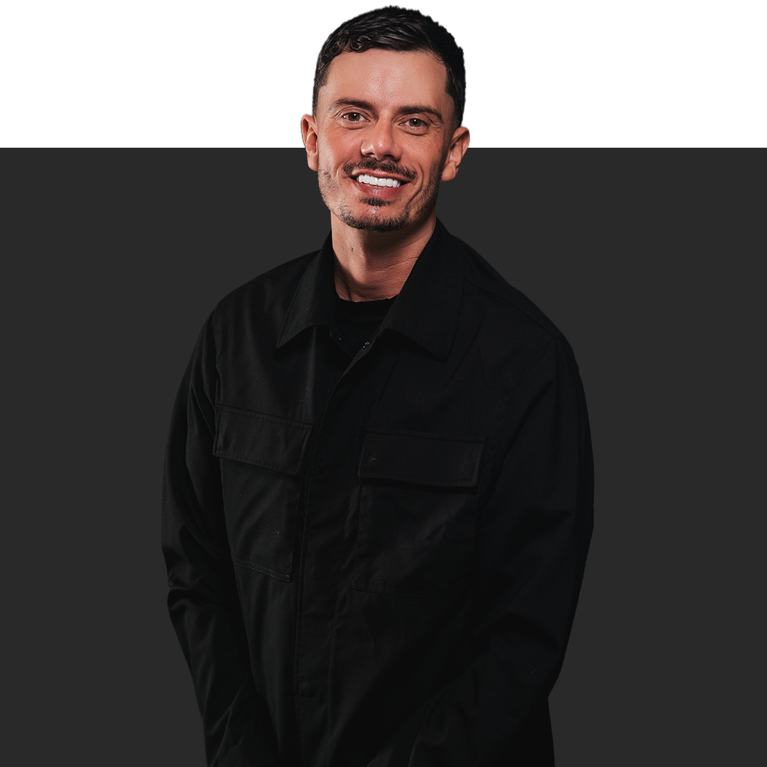 Portret van een man met kort donker haar, lichte huid en een lichte baard, lachend, gekleed in een zwarte jas, tegen een grijze achtergrond.